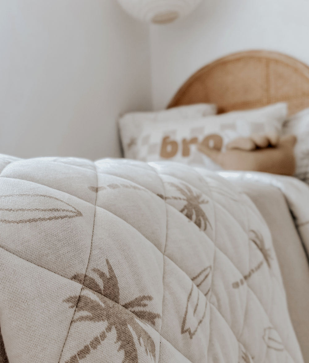 Beige quilt with palm tree patterns on a bed with white pillows and a rattan headboard.