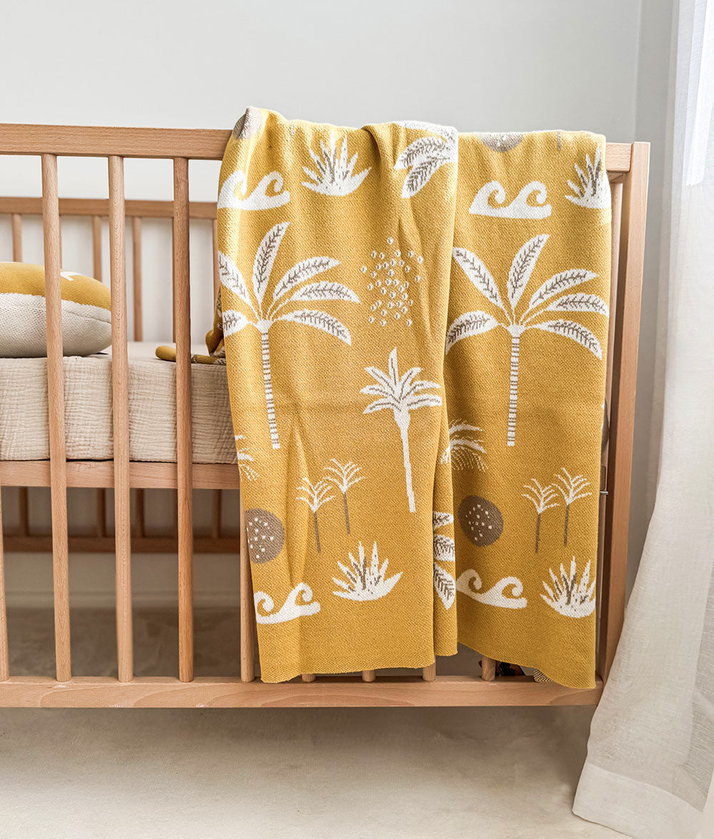 A mustard yellow cotton blanket with a white palm tree pattern draped over a light wood crib in a nursery setting.