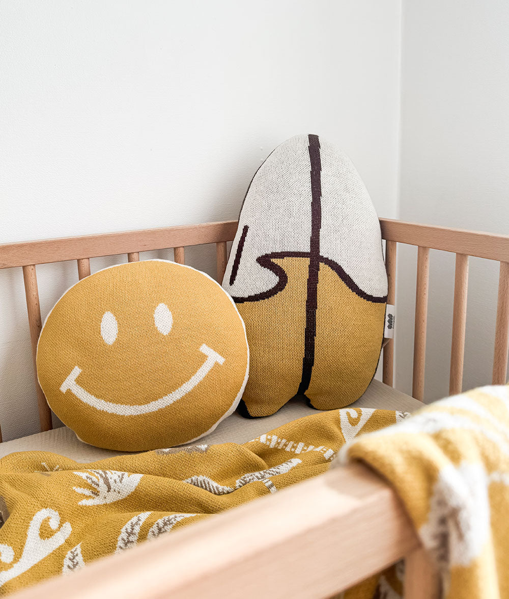 A cozy nursery scene with two textured throw pillows in a wooden crib. One is a round mustard yellow pillow with a white smiley face, and the other is a tall, surfboard-shaped pillow in cream and mustard with dark brown lines.
