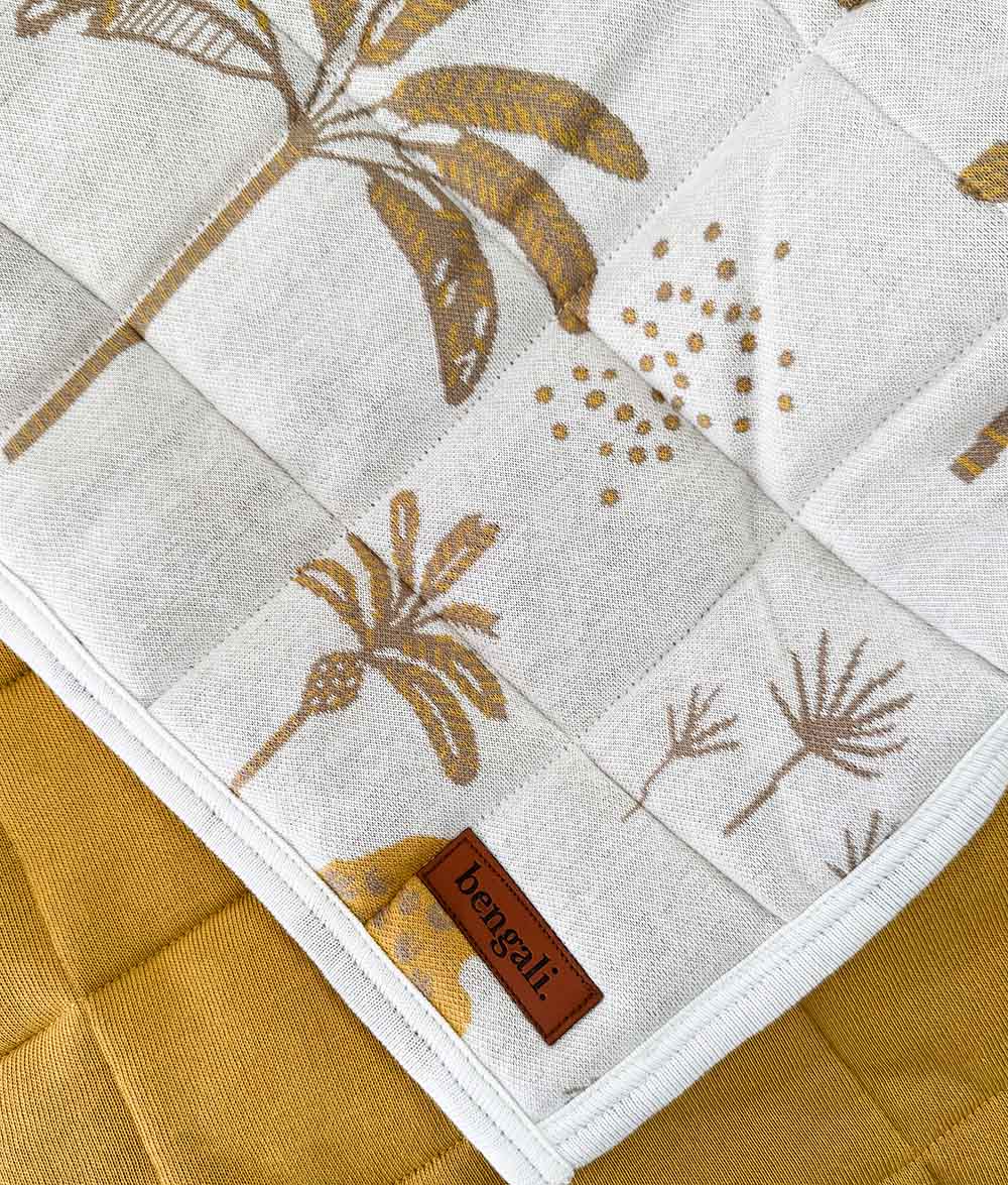 A close-up of a quilted reversible quilt showing a white fabric with a gold and brown palm botanical pattern, layered over a mustard yellow quilted surface. A small brown leather brand tag reading 'bengali' is visible.