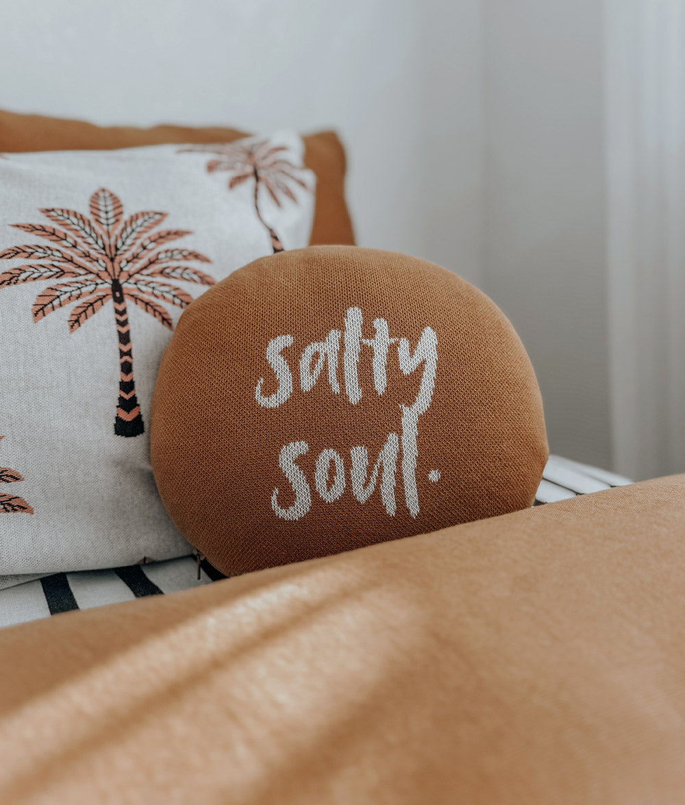 A round tan throw pillow with 'salty soul.' embroidered in white script, resting in front of a white pillow with palm tree embroidery on a bed.