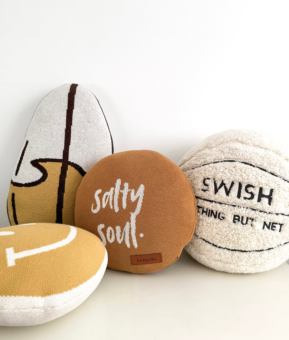 Four novelty beach-themed plush pillows arranged together on a white surface against a white background. One pillow is tobacco tan with the words 'salty soul'.