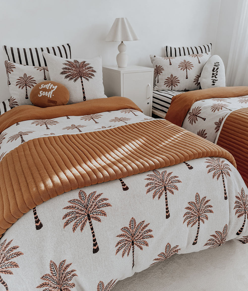 A styled bedroom scene showing a bed dressed in a white duvet cover and pillow shams printed with brown palm trees, plus a rust-orange ribbed throw blanket and matching accent pillows. A second bed is partially visible in the background, along with a white nightstand and lamp.