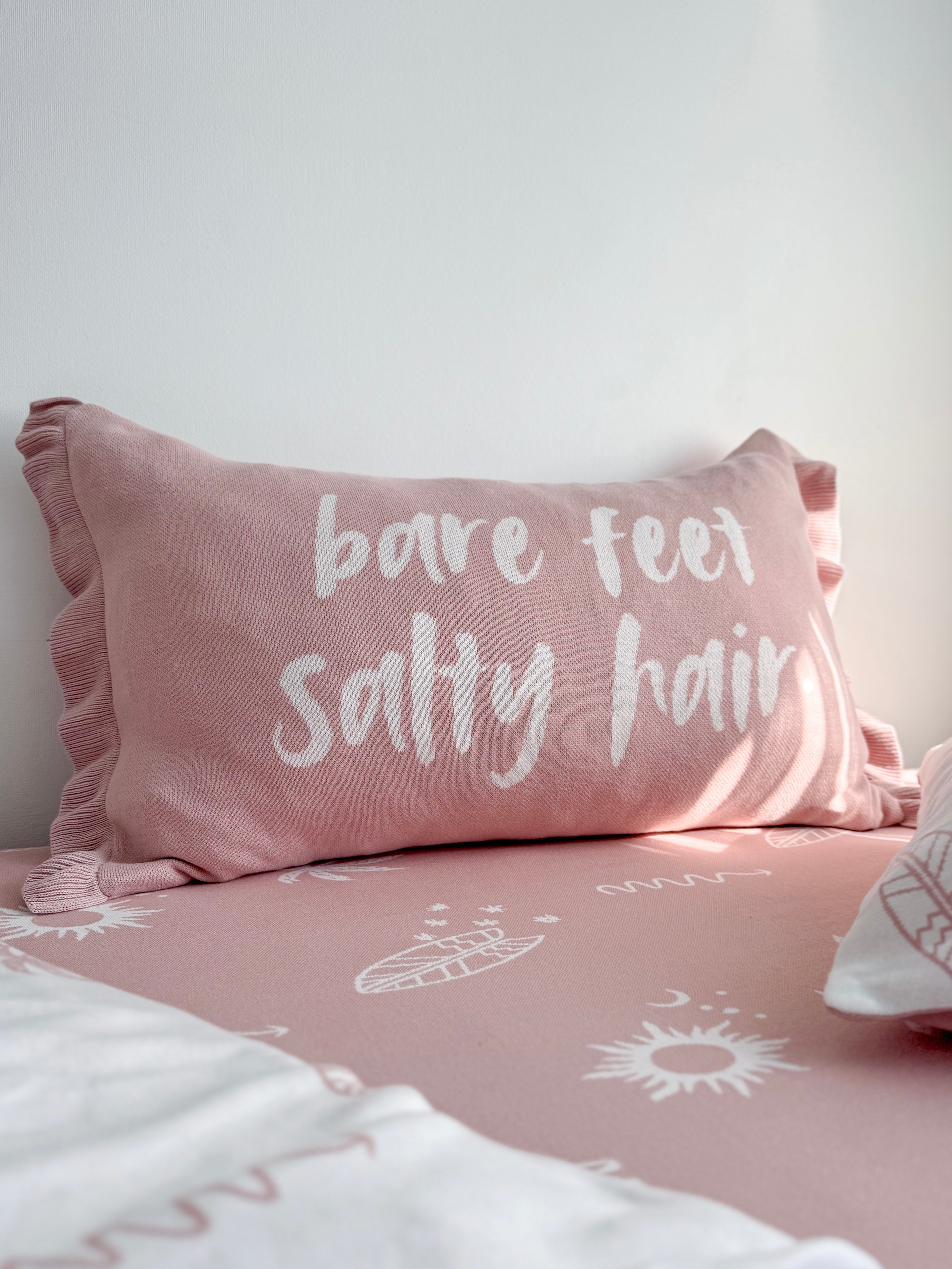 A pink bedding set with a matching pillow featuring the white embroidered text 'bare feet salty hair', shown on a bed against a light gray wall.