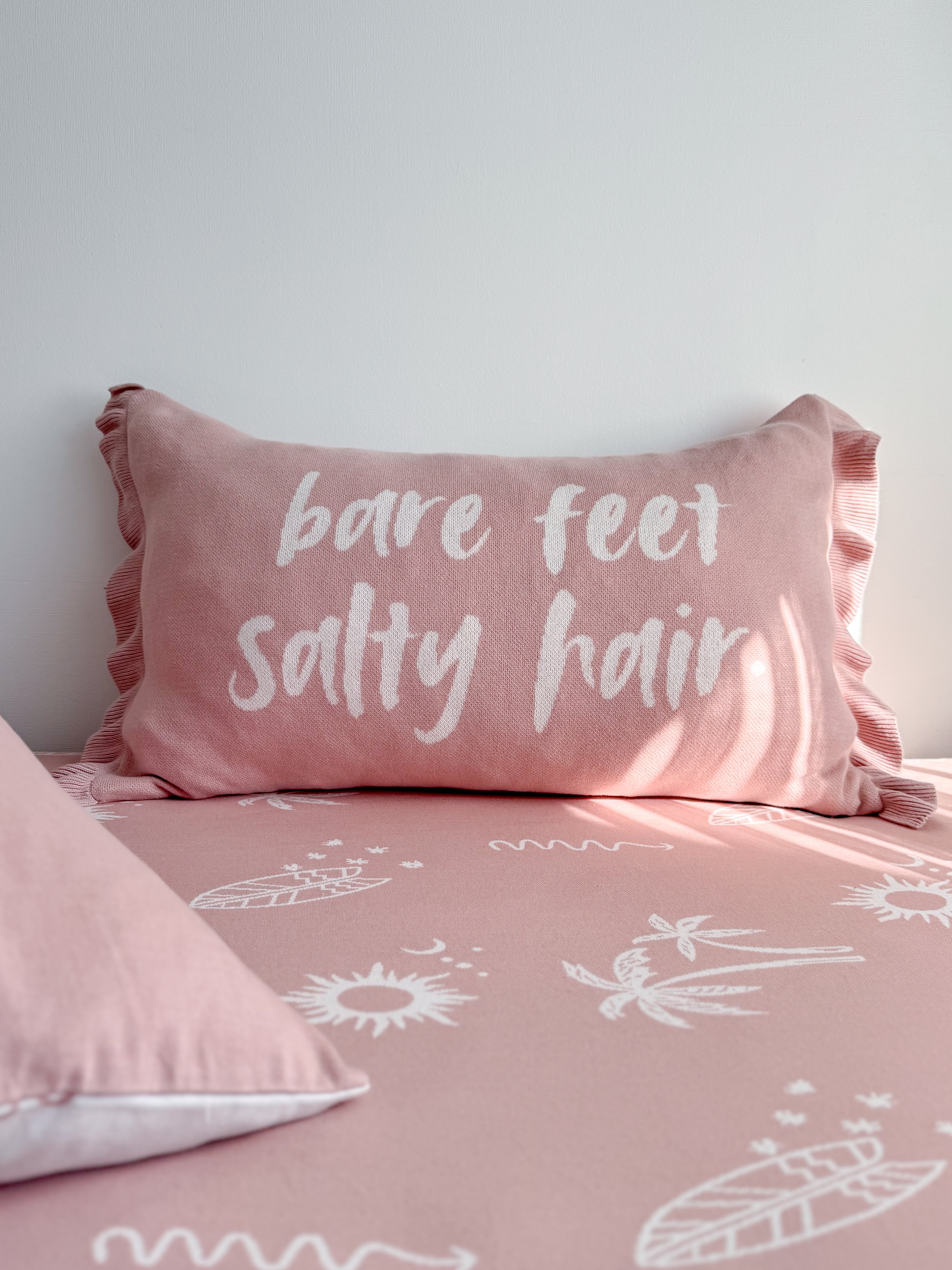 A pink bedding set with a matching pillow featuring the white embroidered text 'bare feet salty hair', shown on a bed against a light gray wall.