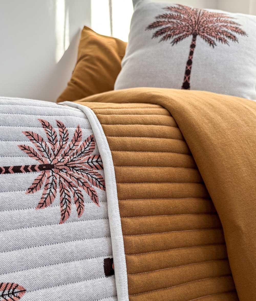 A close-up of a bed styled with a white quilted throw blanket featuring pink and brown palm tree embroidery, a folded mustard yellow ribbed blanket, and matching pillows.