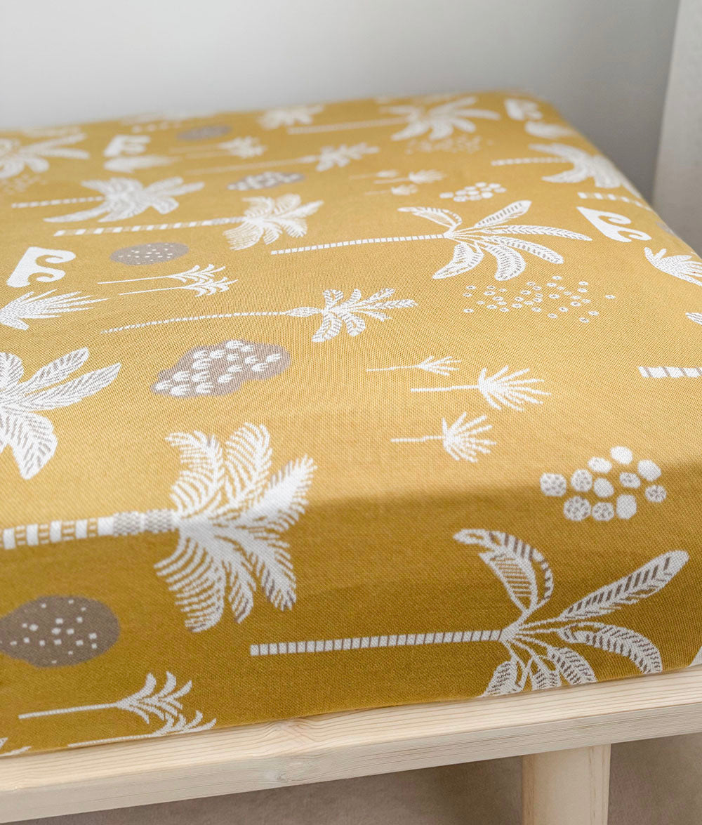 A mustard yellow fitted jersey cotton sheet with a white and gray tropical palm and wave pattern, resting on a light wood bench.