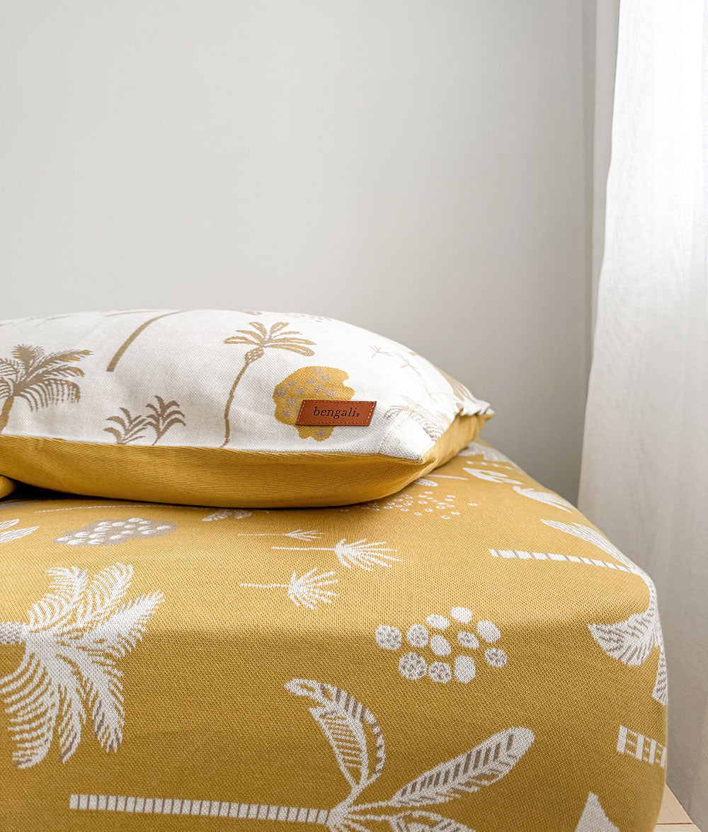 A close-up of a yellow and white tropical patterned jersey cotton sheet, with a matching mustard yellow Golden Hour pillowcase featuring a palm tree design and a small 'bengali' brand tag.