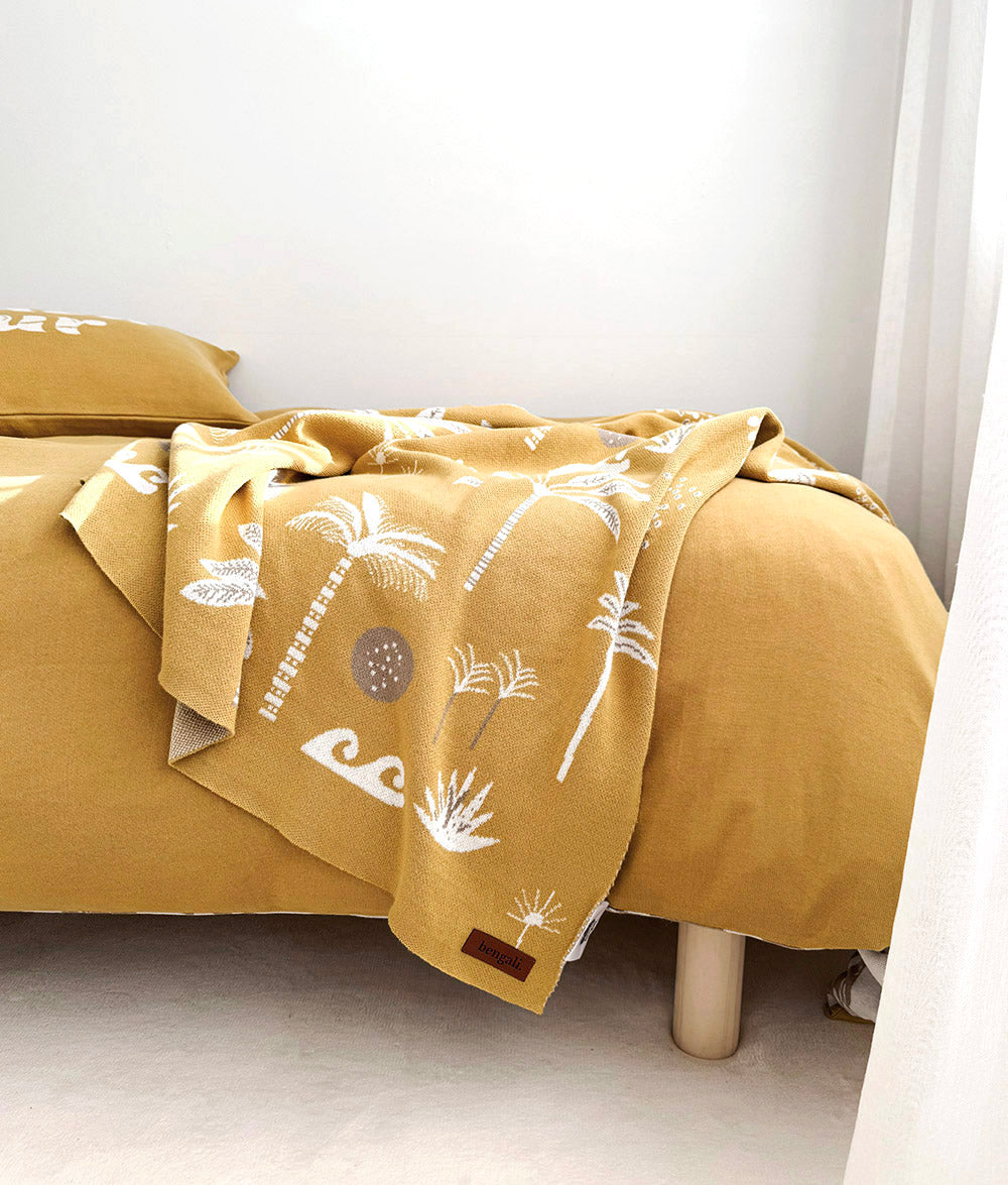 A mustard yellow duvet cover and pillowcase set with a white palm tree and wave pattern, styled on a bed in a minimalist room.