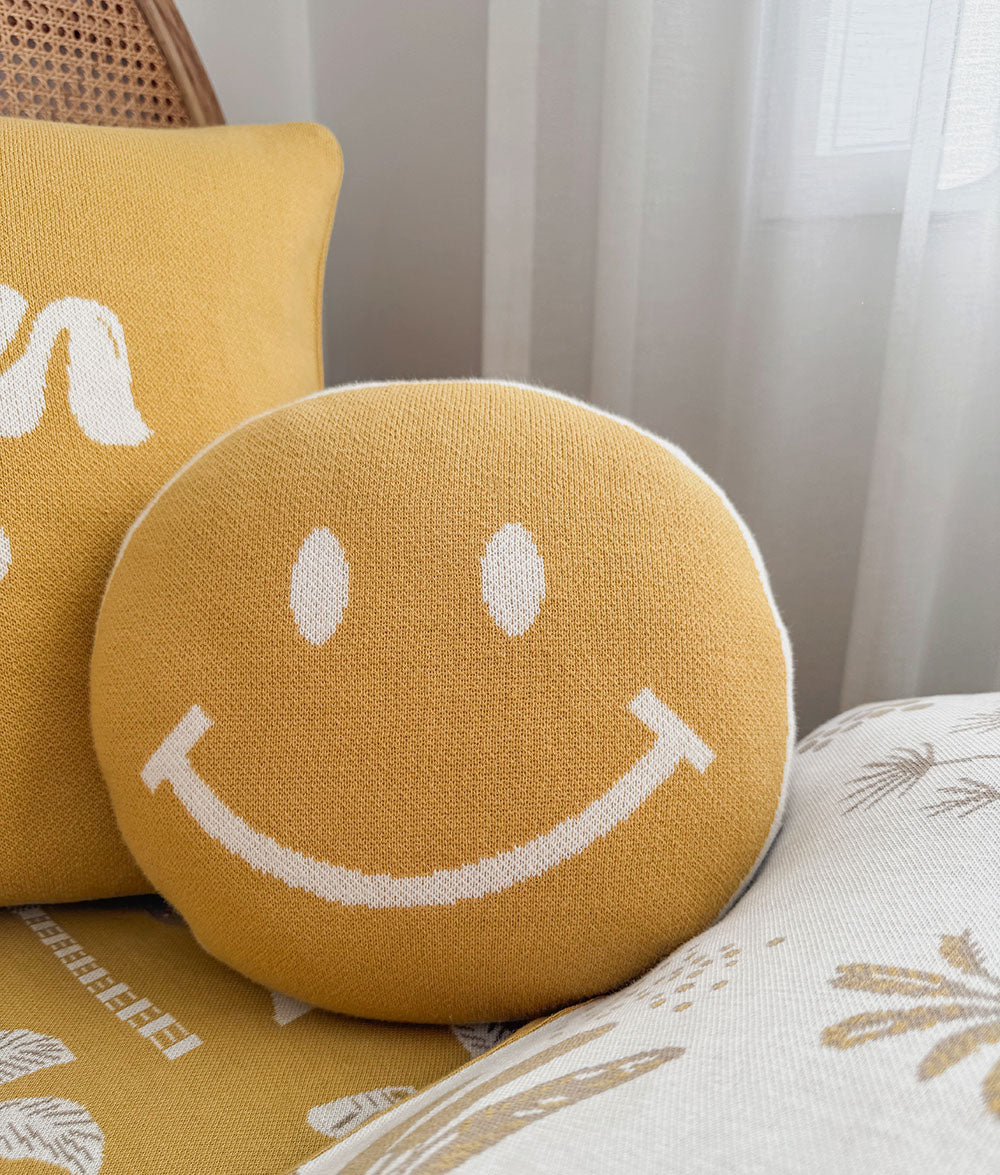 A round yellow throw pillow with a white knit smiley face sits on a patterned sofa next to another yellow pillow in a bright room.