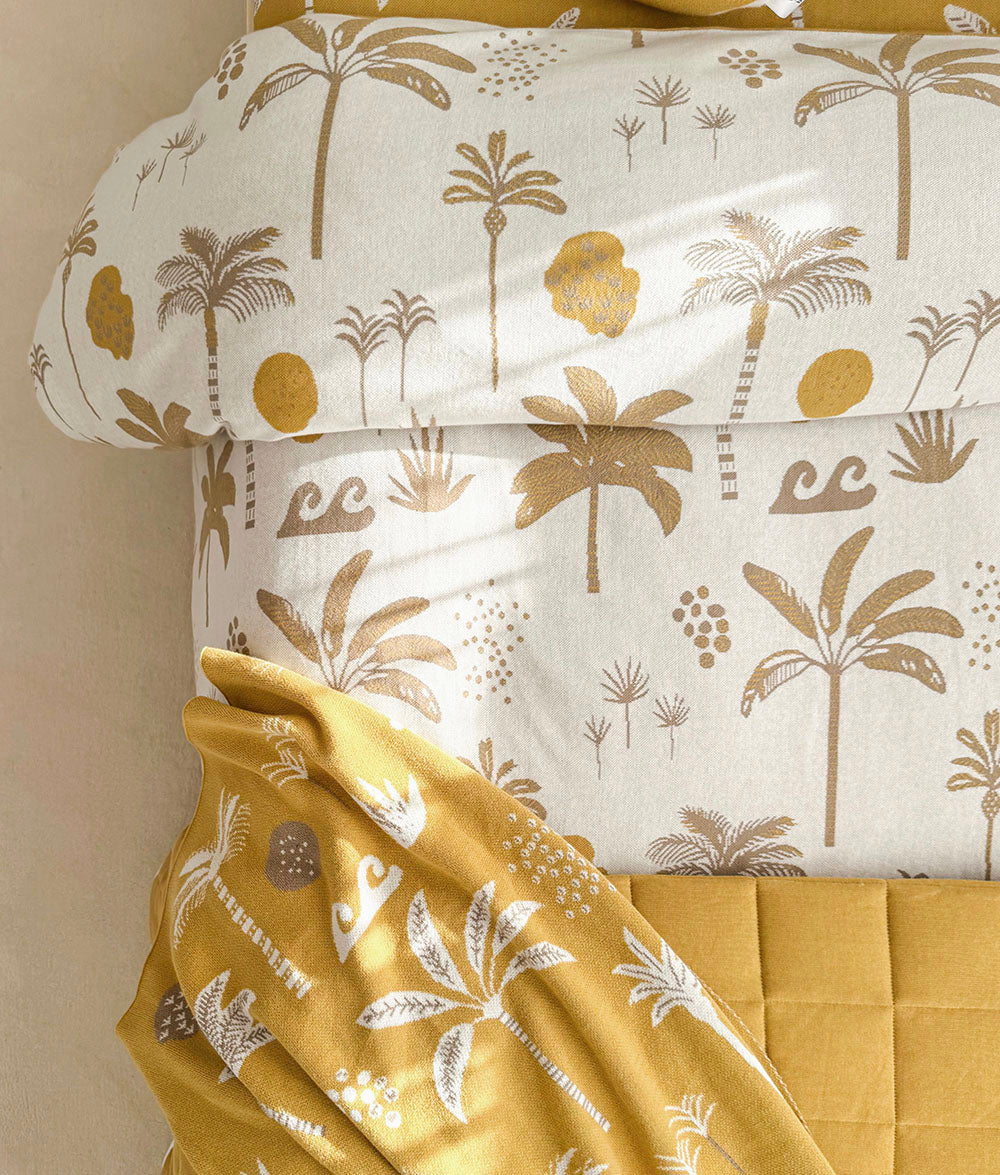 A close-up of the Golden Hour bedding set featuring a duvet cover and matching pillowcase with a gold palm tree and sun motif on an off-white background, styled with a mustard yellow quilted blanket.