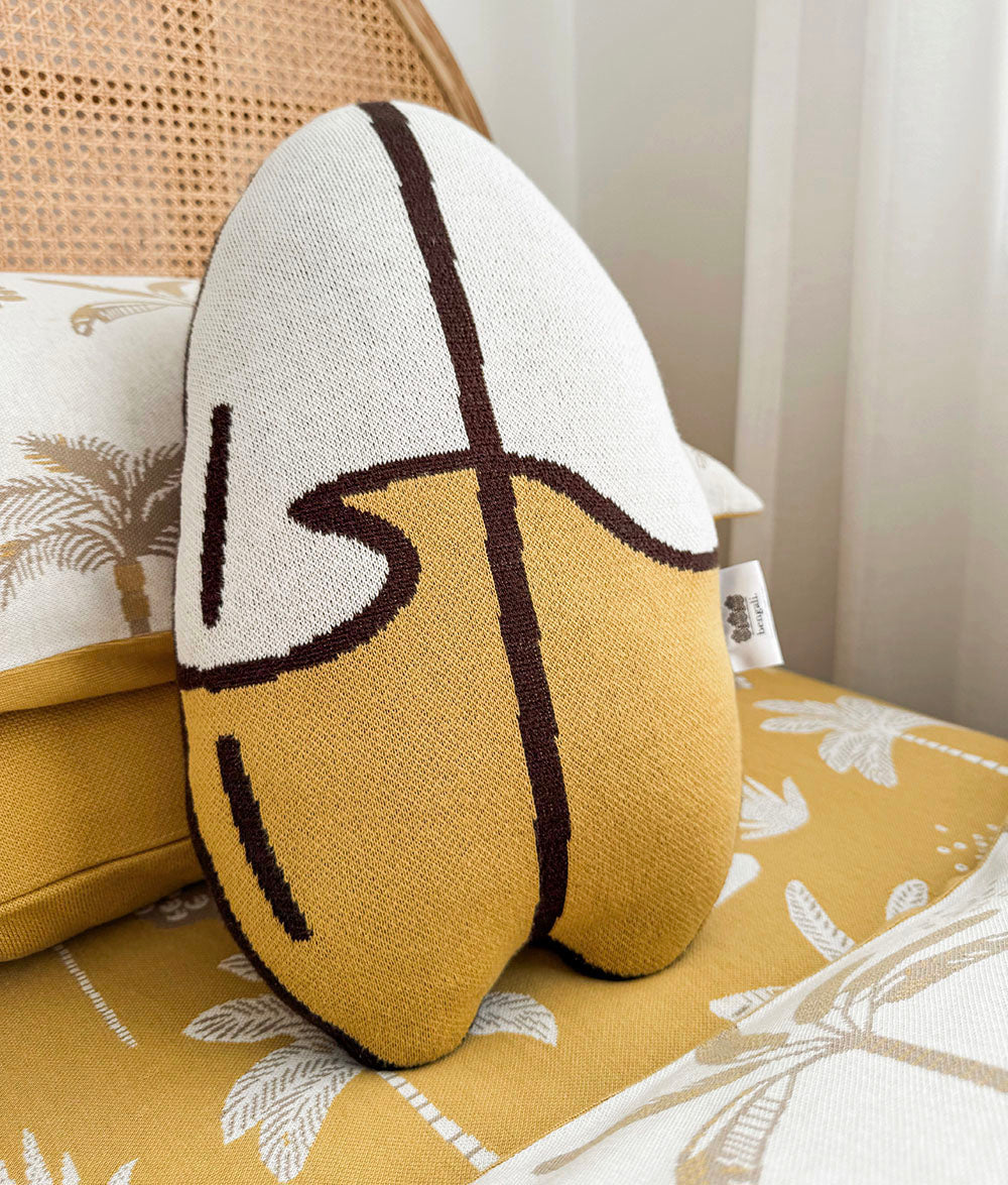 A woven surfboard-shaped throw pillow in mustard yellow and off-white with dark brown abstract lines, styled on a patterned single bed.