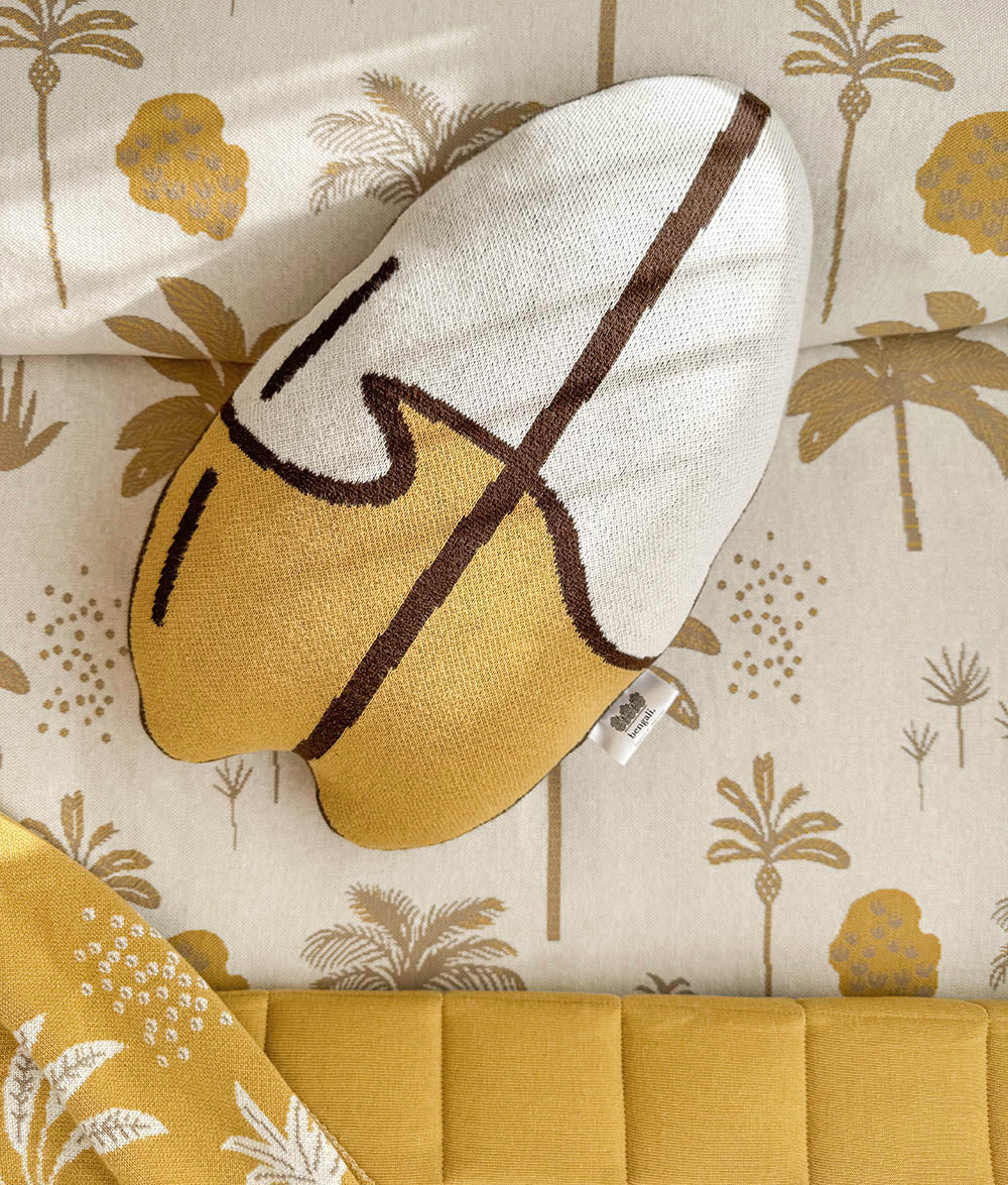 A knitted woven surfboard cushion in a cream and mustard yellow colourway with brown abstract lines, styled on a matching palm patterned duvet cover.