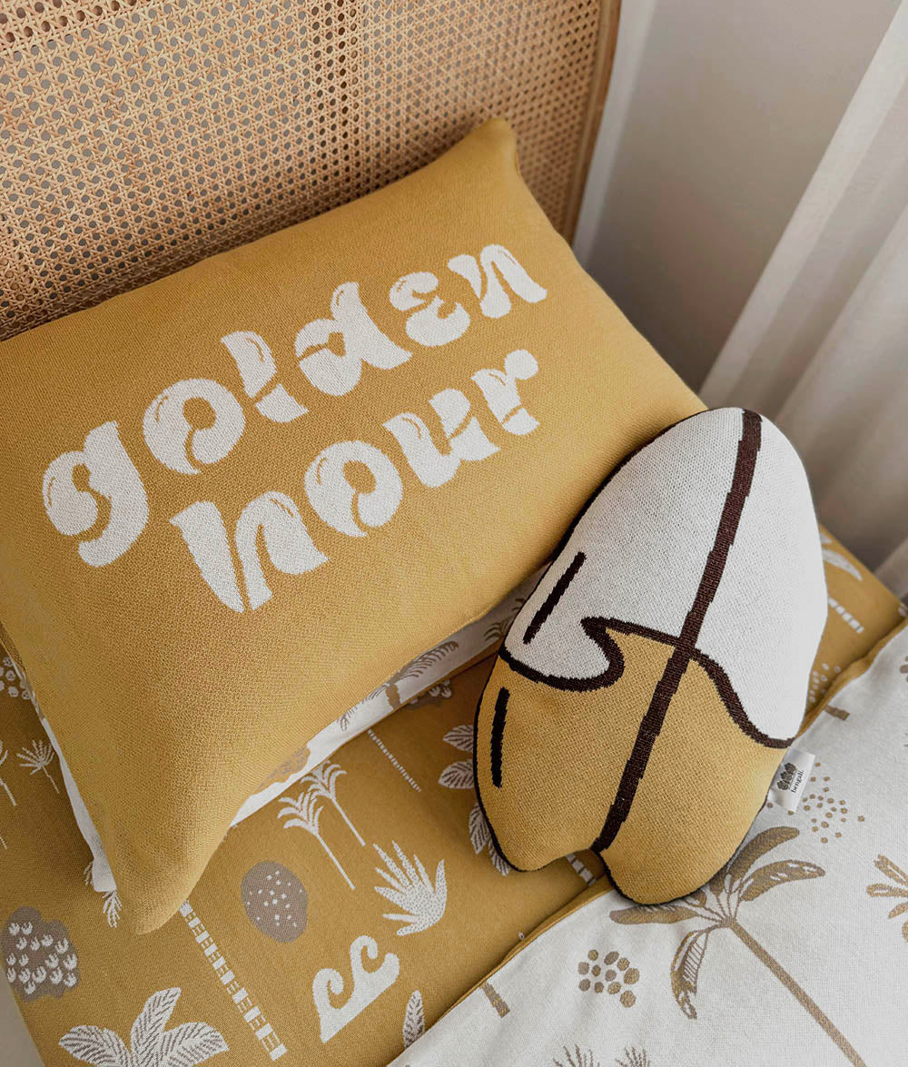 A mustard yellow pillowcase with 'golden hour' written in white script, styled on a cane-back headboard next to a patterned cushion and a white accent surfboard shaped pillow.