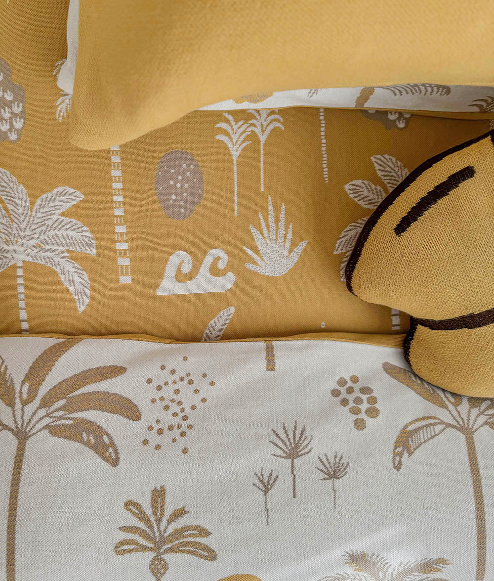 A close-up of a bedding set featuring a mustard yellow sheet and pillowcase with white palm tree and botanical patterns, paired with a white duvet cover and pillow in a taupe brown botanical print, styled with a yellow and brown accent pillow.