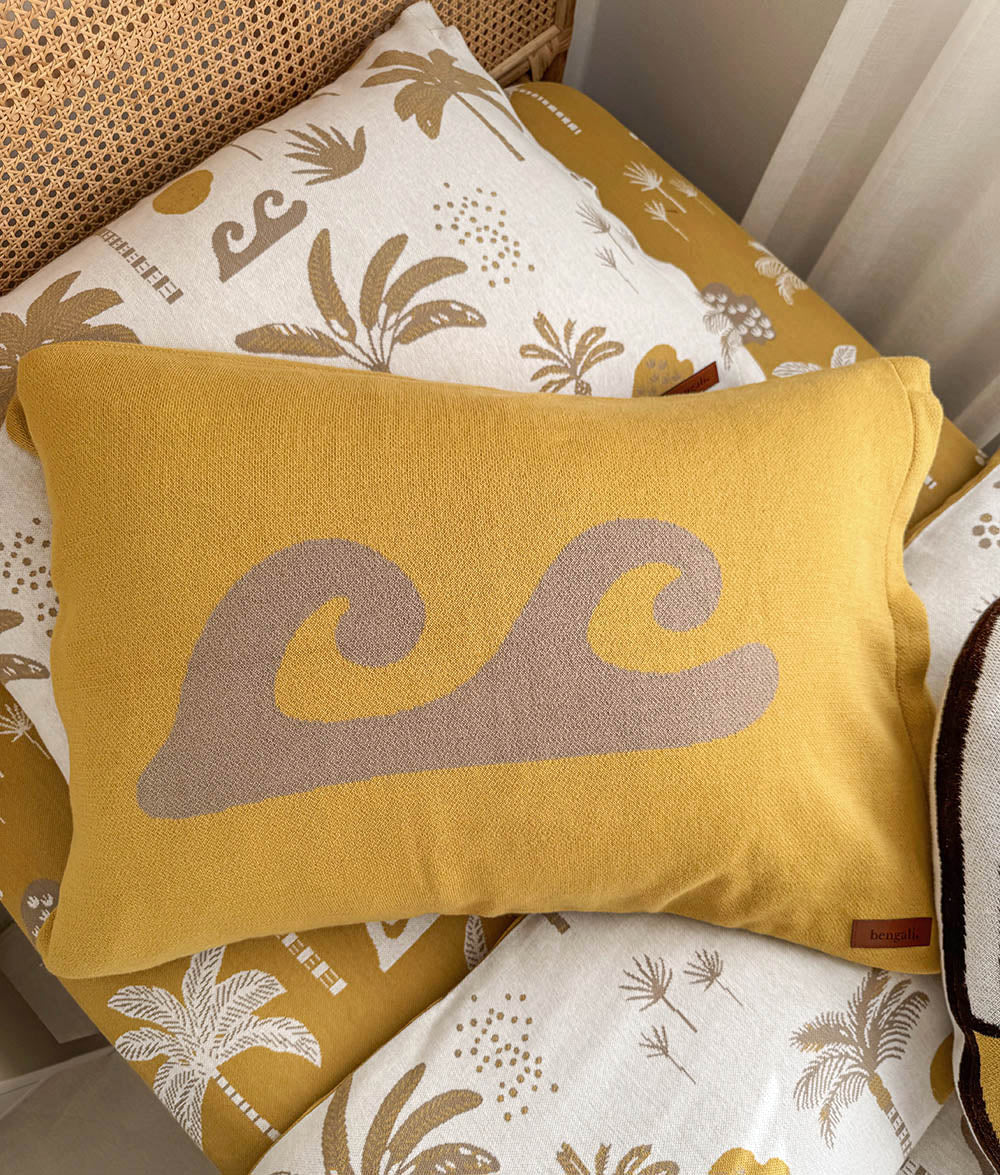 A yellow rectangular throw pillow with a large taupe wave design, resting on a bed with tropical patterned sheets.