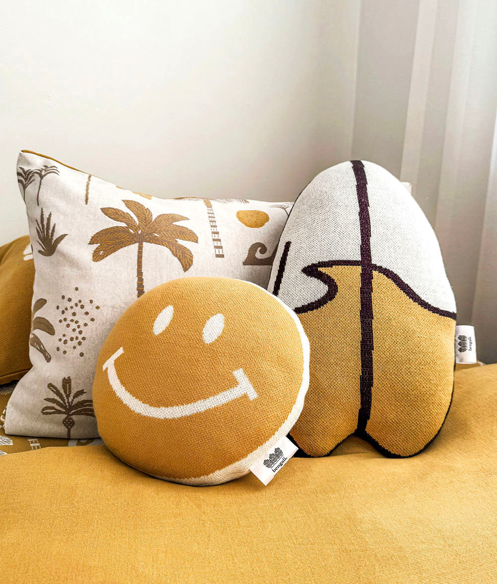 Three decorative throw pillows arranged on a mustard-yellow upholstered surface against a light neutral wall. The front pillow is round and tan with a white smiley face.