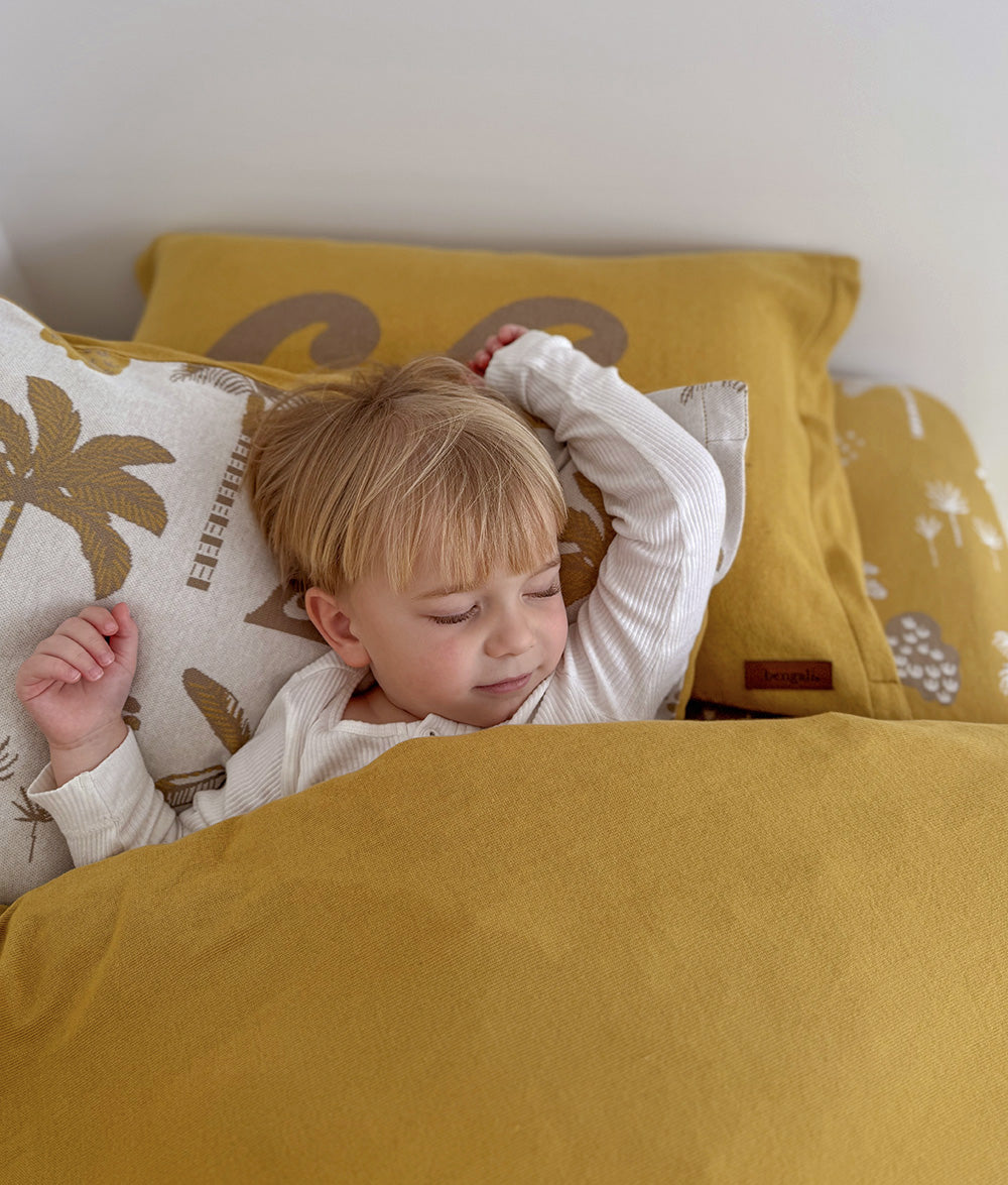 A young child sleeps peacefully under the Golden Hour jersey cotton duvet cover in warm mustard. The bed is styled with matching Golden Hour pillowcases featuring soft taupe wave motifs and palm tree prints on a natural base, creating a calm, sun-soaked bedroom with warm coastal tones.