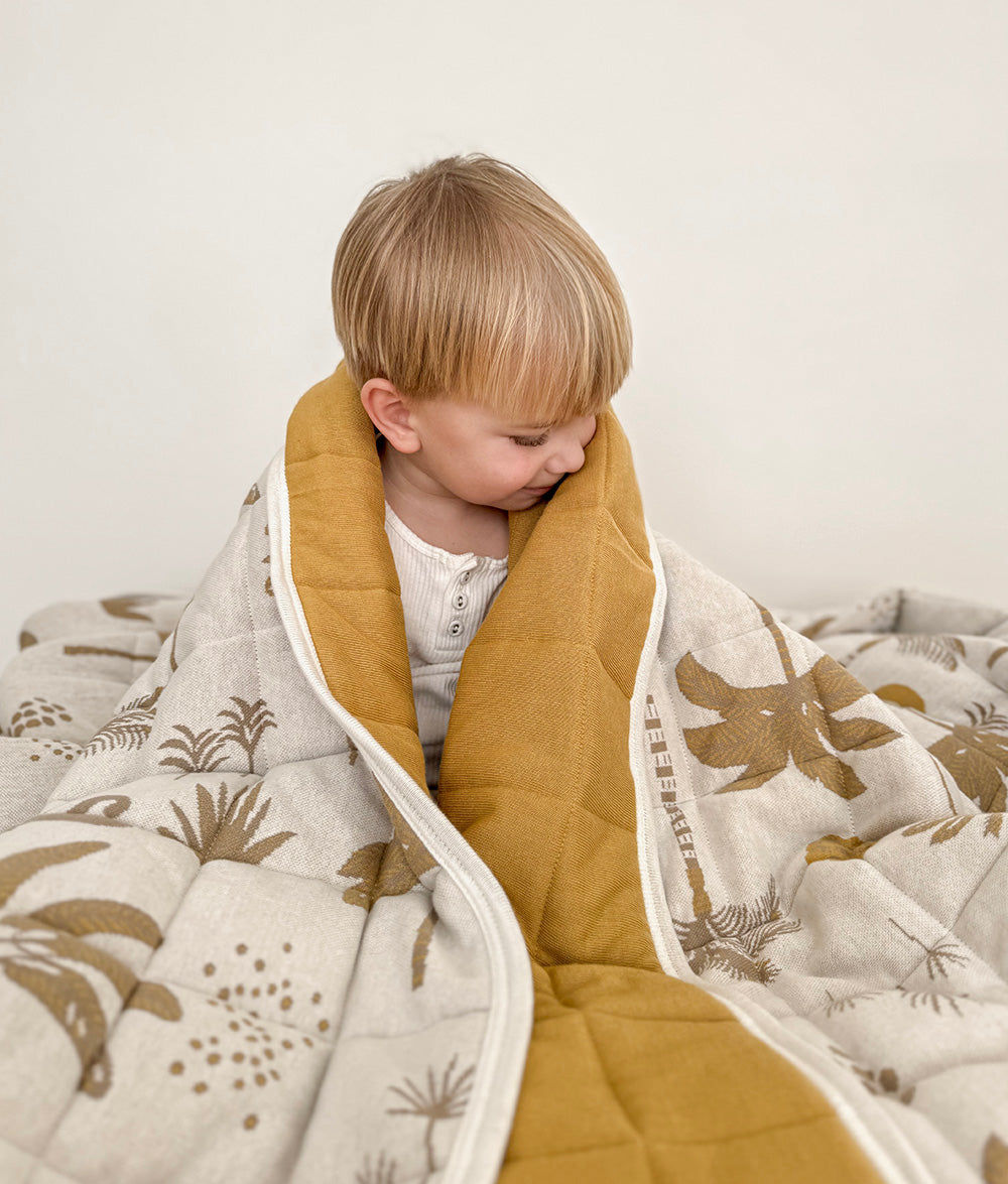 Child wrapped in the Golden Hour reversible quilt, showcasing the plain golden mustard reverse and ivory coastal print front.