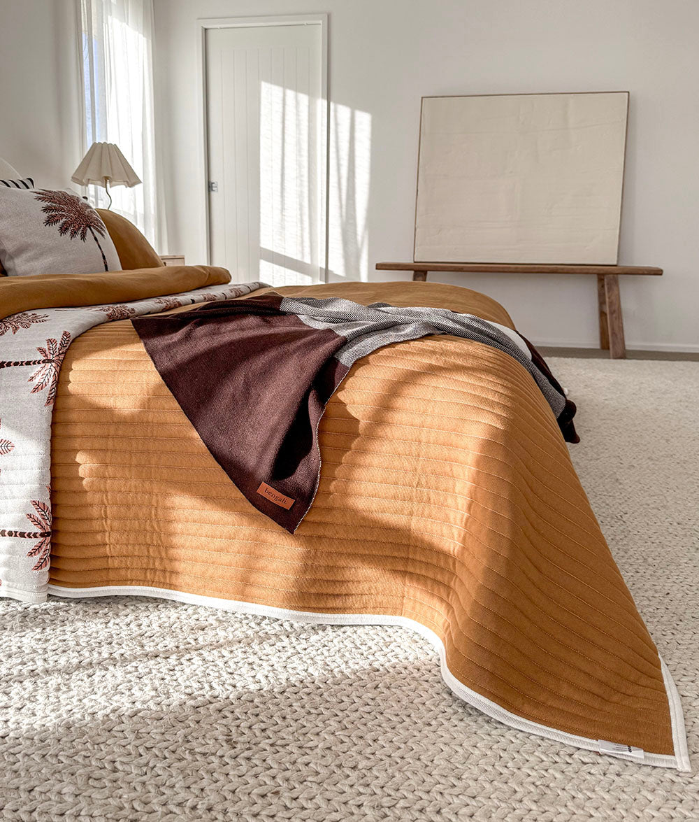 A quilted tobacoo bedding set styled on a bed in a bright, modern bedroom. The set includes a ribbed quilt with a white border and a matching quilted pillowcase with a botanical pattern.