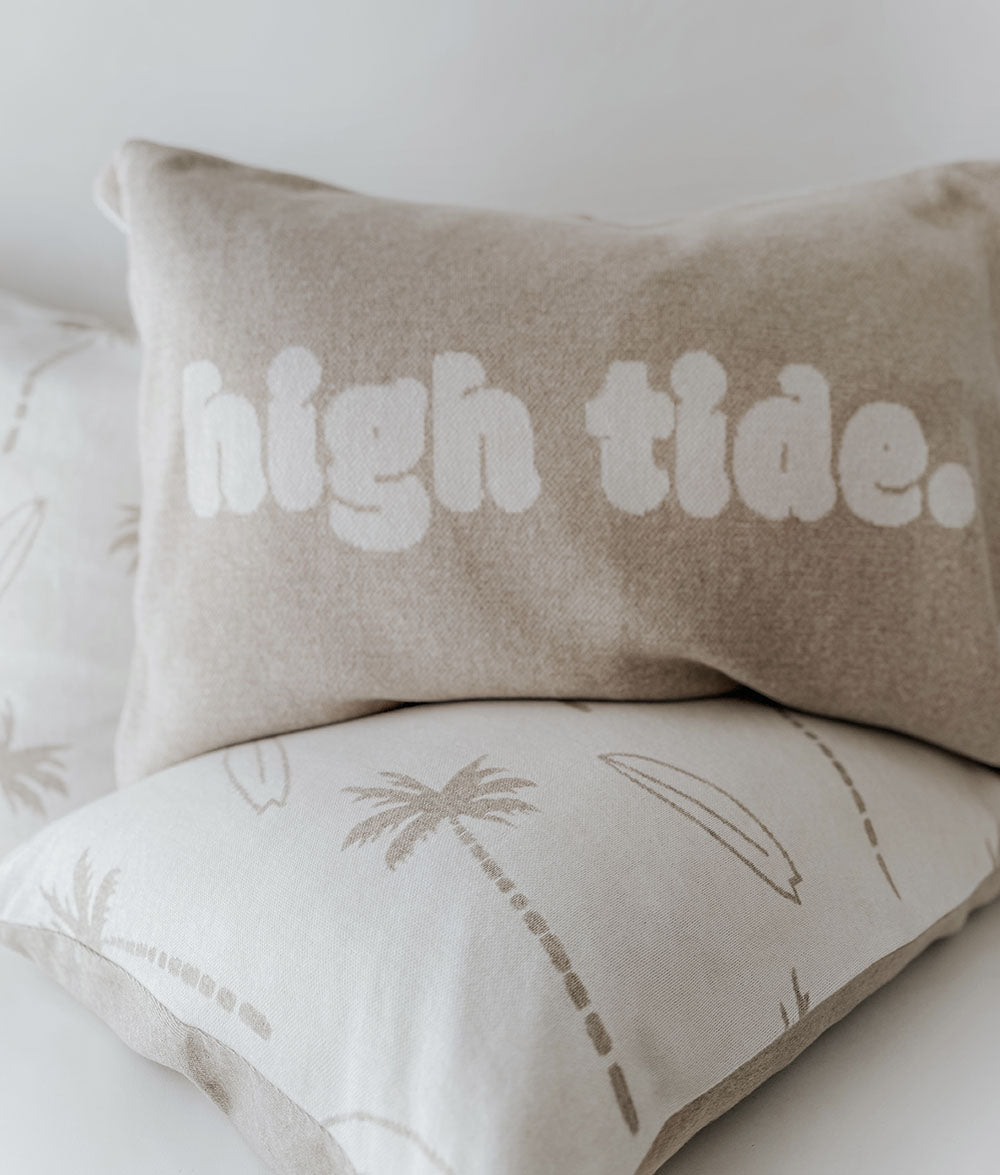 Beige pillow with 'high tide.' text on a bed with palm tree and surfboard design.