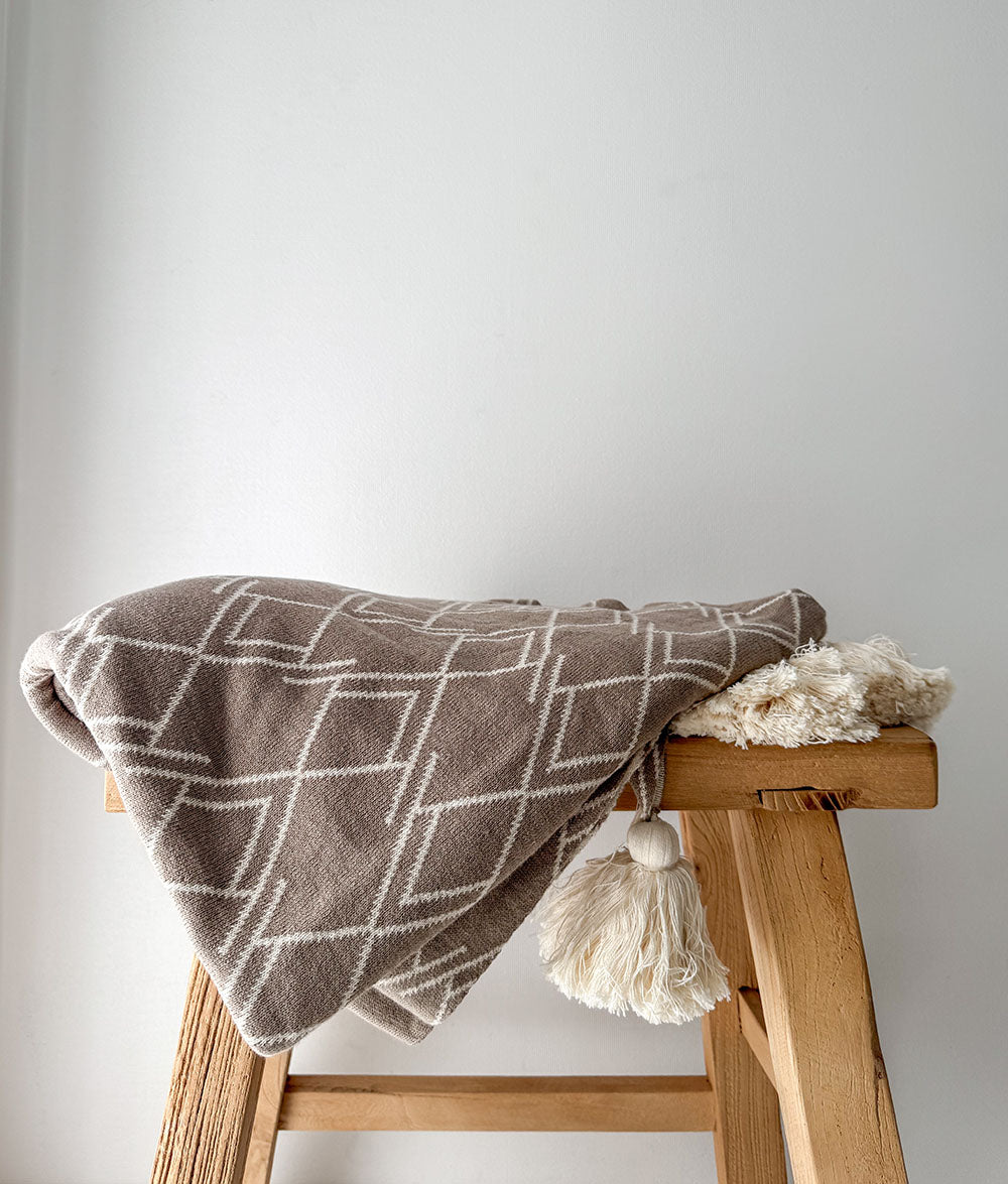 Brown patterned blanket with tassels draped over a wooden stool against a light gray background