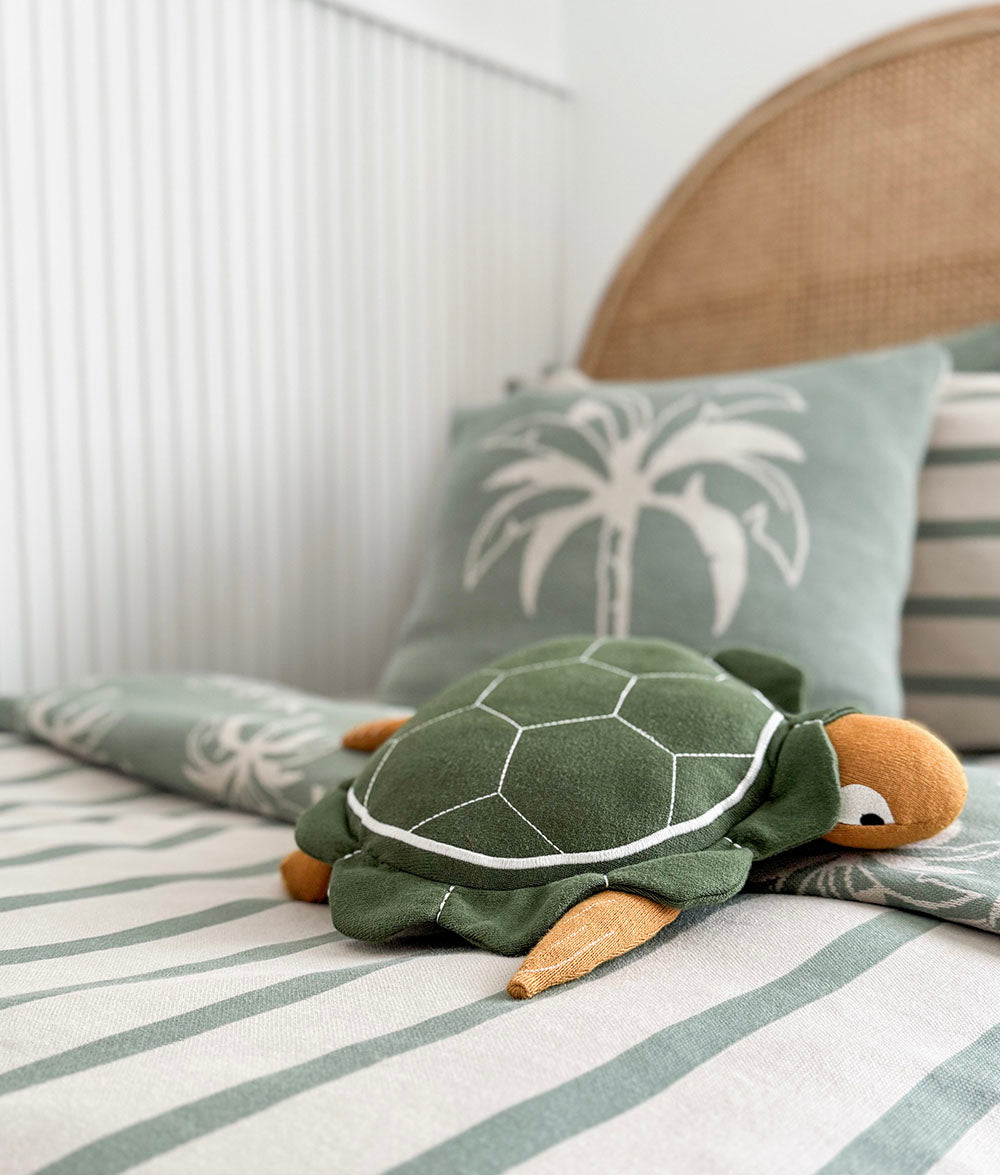 Green plush turtle toy on a bed with striped bedding and decorative pillows.