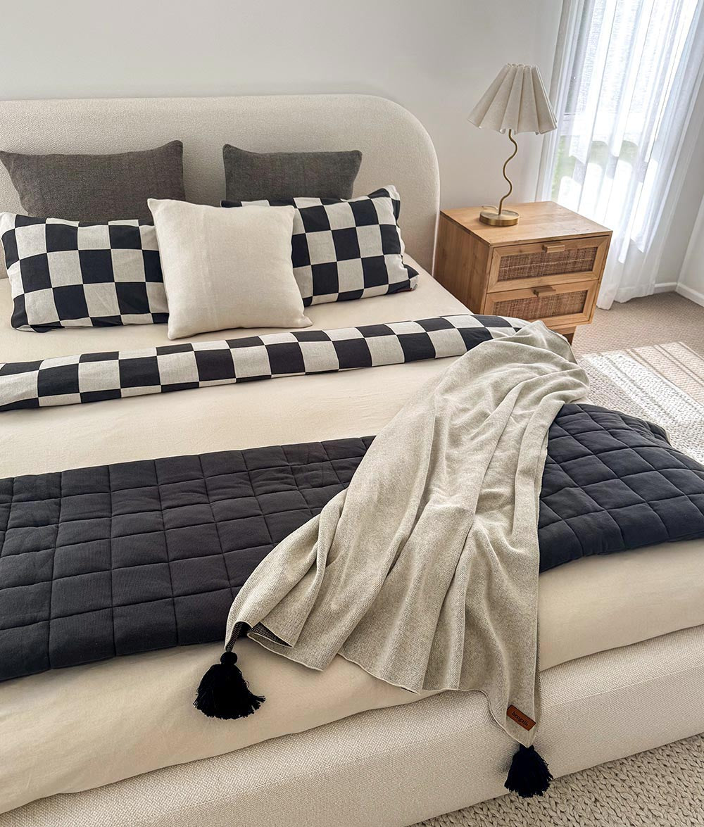 Neatly made bed with checkered and solid pillows, a blanket, and a lamp on a nightstand.