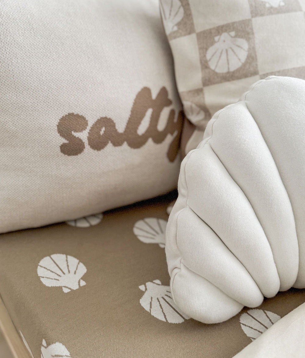 Close-up of a cushion with 'salty' text and shell patterns on a beige background.