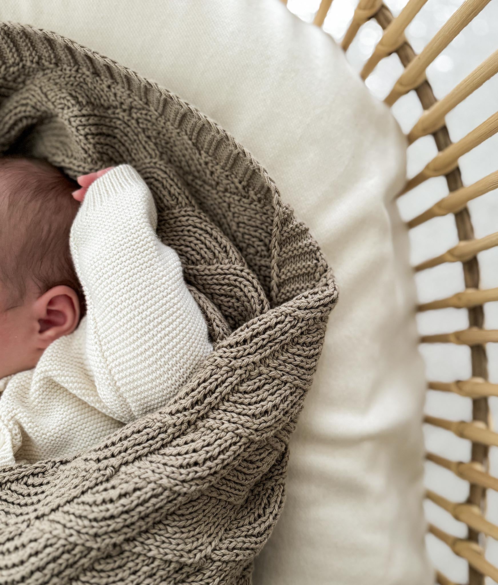 Newborn baby wrapped in a brown knitted blanket on a white pillow with a rattan basket in the background.