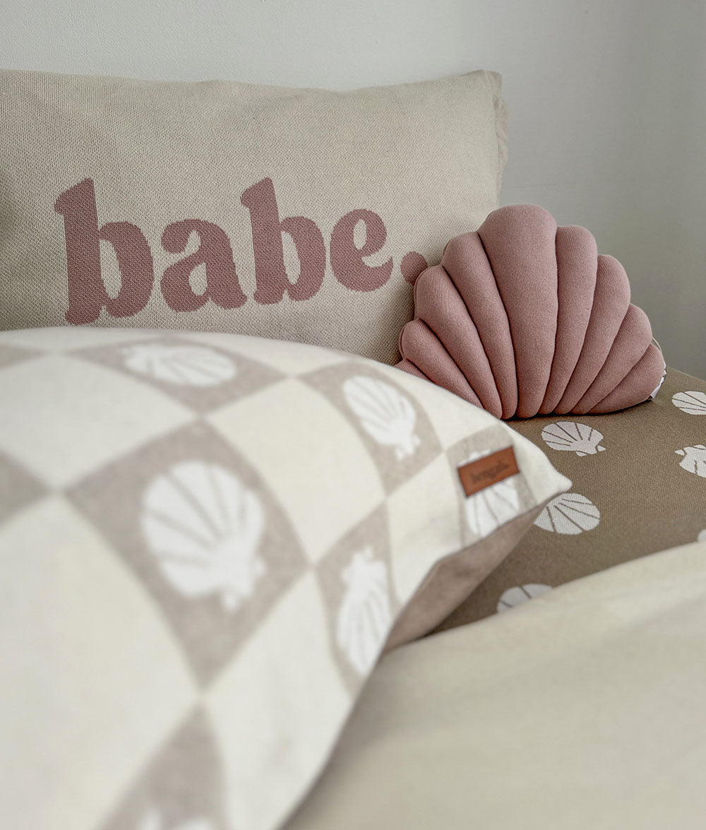 Decorative pillow with 'babe' text and a shell-shaped pillow on a bed with a shell-patterned blanket.