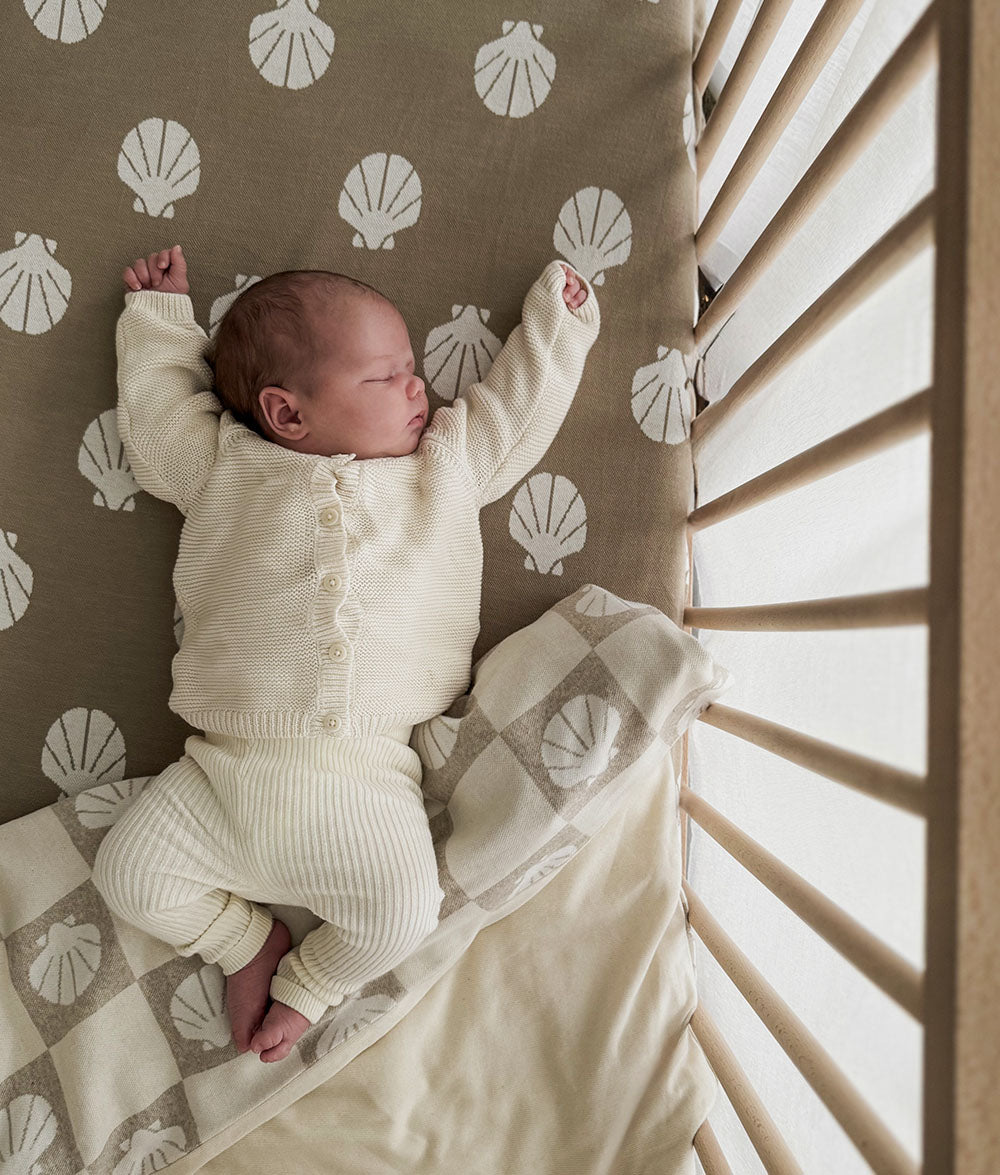 Newborn baby in a white outfit lying on a crib with shell-patterned bedding.