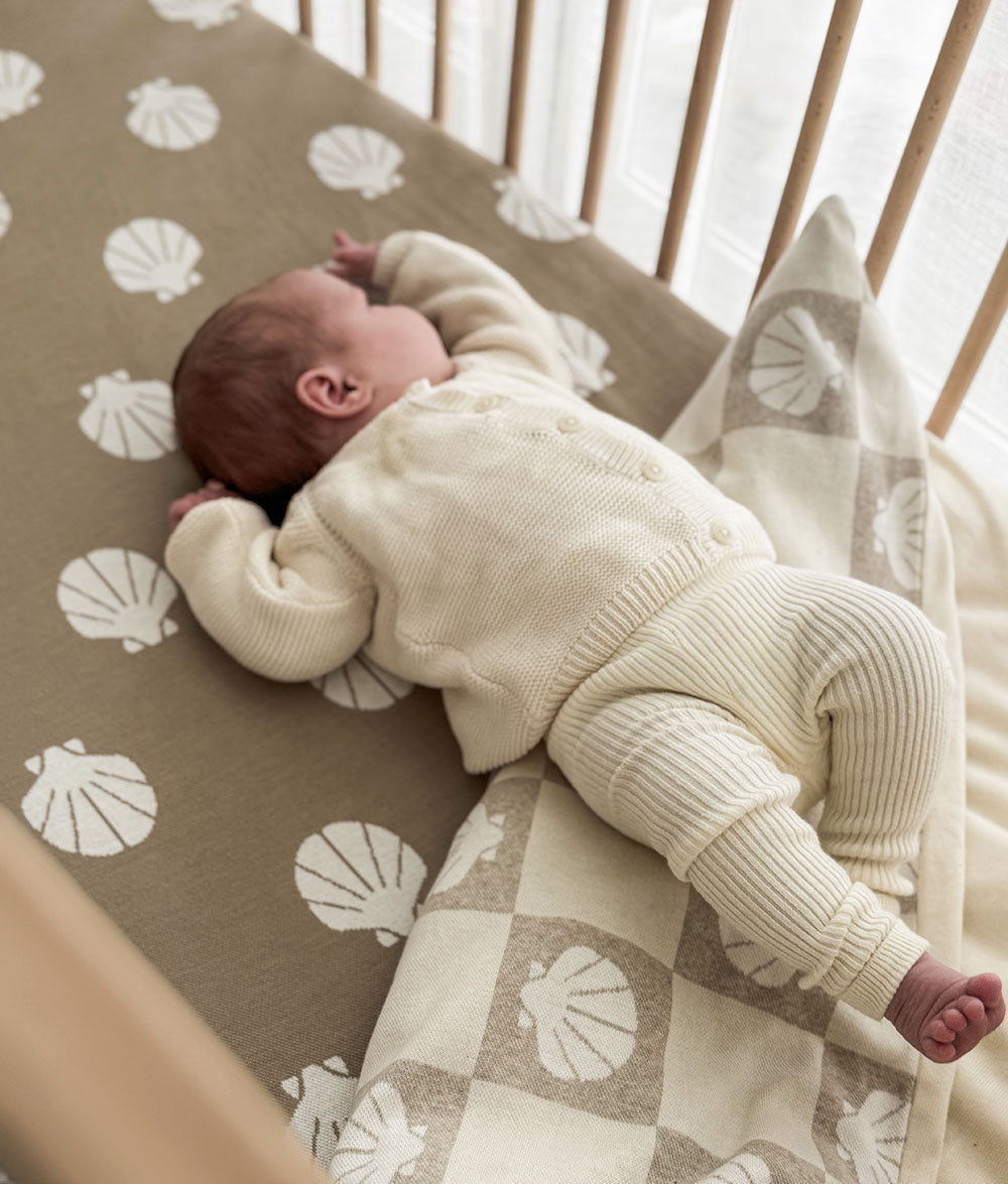 Newborn baby in a cream outfit lying on a patterned blanket in a crib.