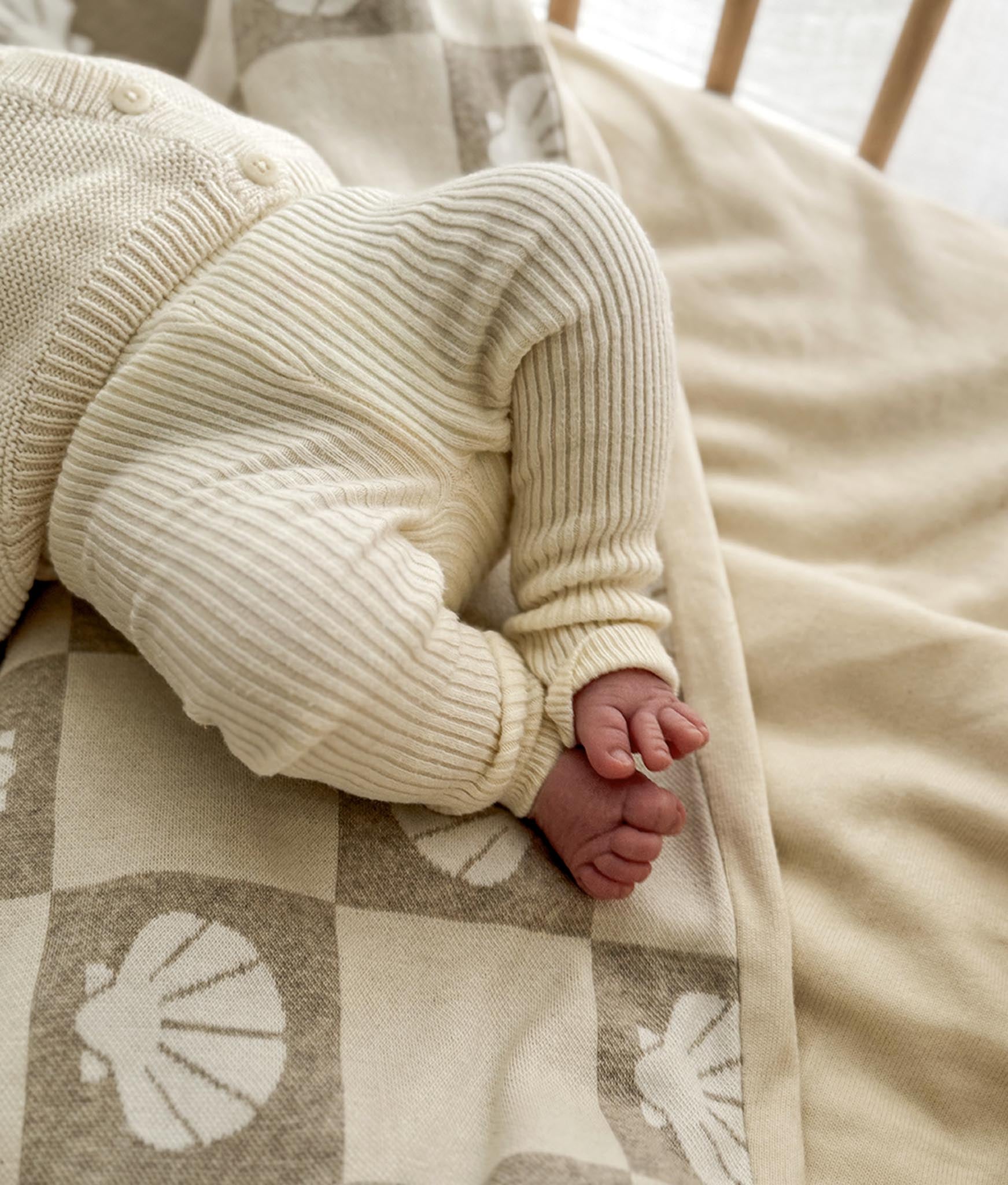 Close-up of a baby's hand and foot wearing a cream knitted outfit on a patterned blanket.