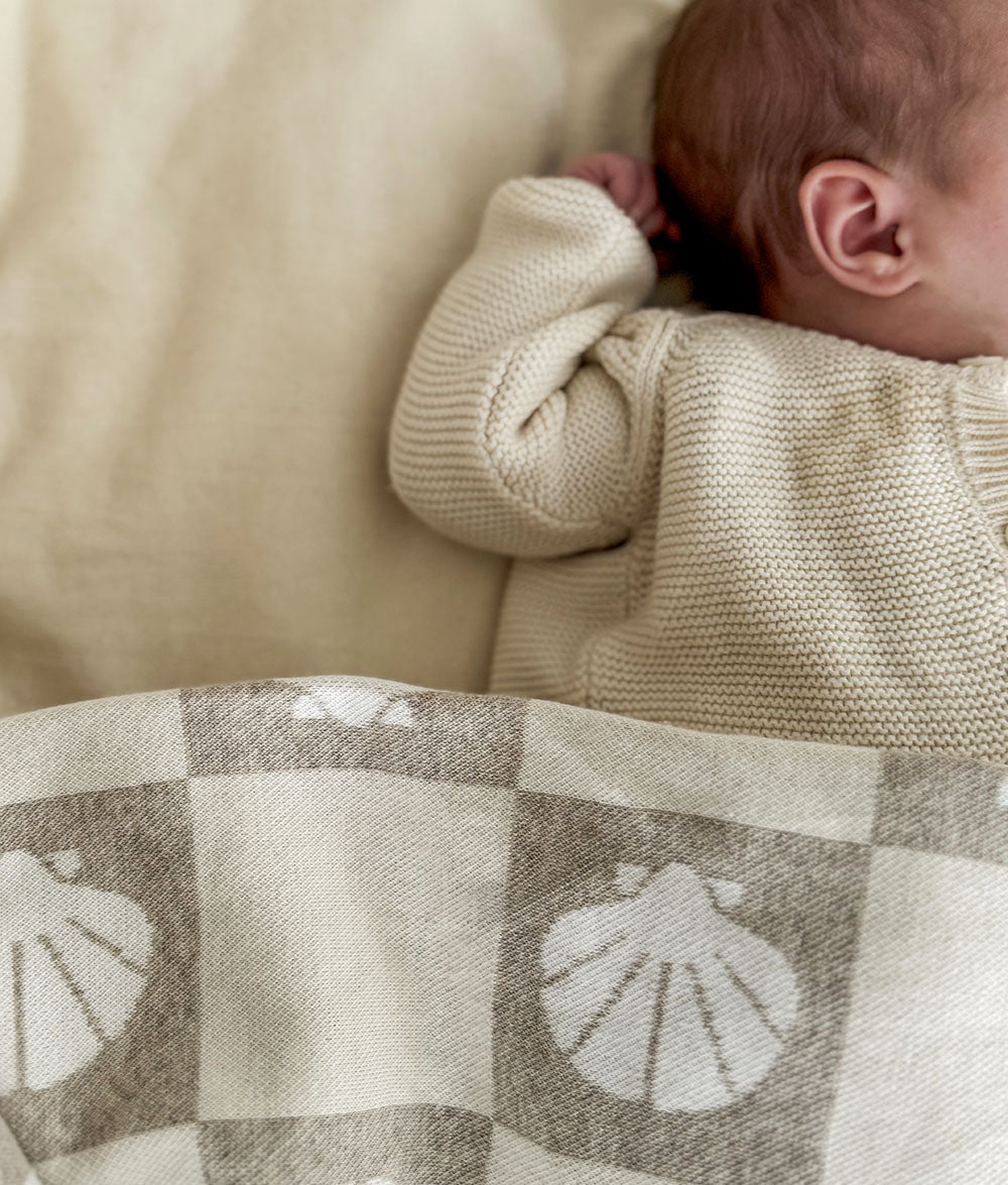 Baby in a beige knit sweater lying on a checkered blanket with shell patterns