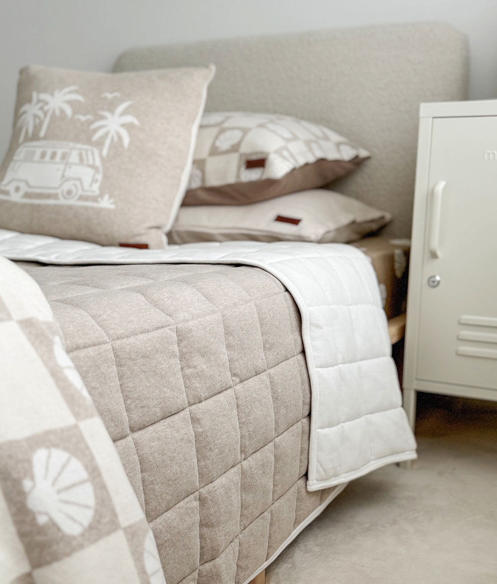 Neatly made bed with beige and white quilt, pillows, and a cabinet in the corner.