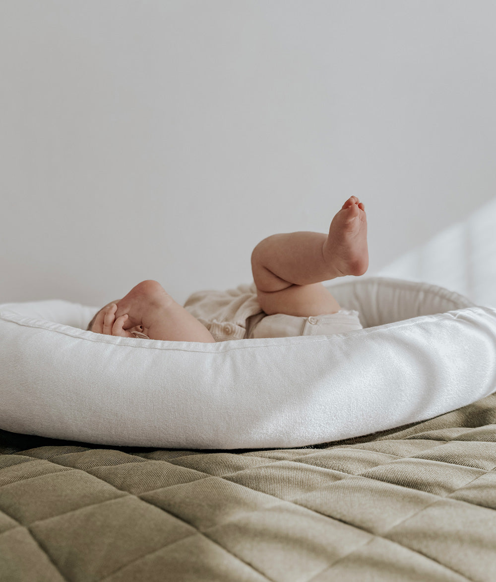 A baby's bare legs kicked in the air while lying inside a white fabric baby lounger bed placed on a quilted beige blanket against a neutral light gray background.