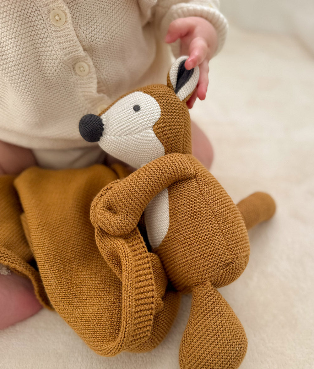 A close-up of a baby in a cream knit cardigan holding a knitted plush fox toy in a warm mustard-brown color, set against a soft cream background.