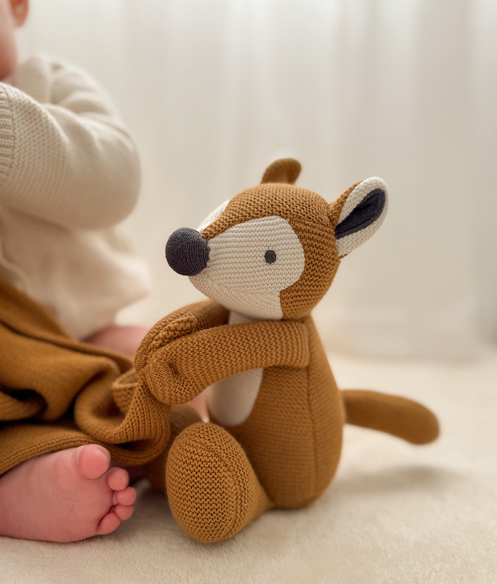 A close-up lifestyle shot of a baby's foot and leg resting on a cozy cream knit blanket next to a knitted plush toy deer in a warm brown color.