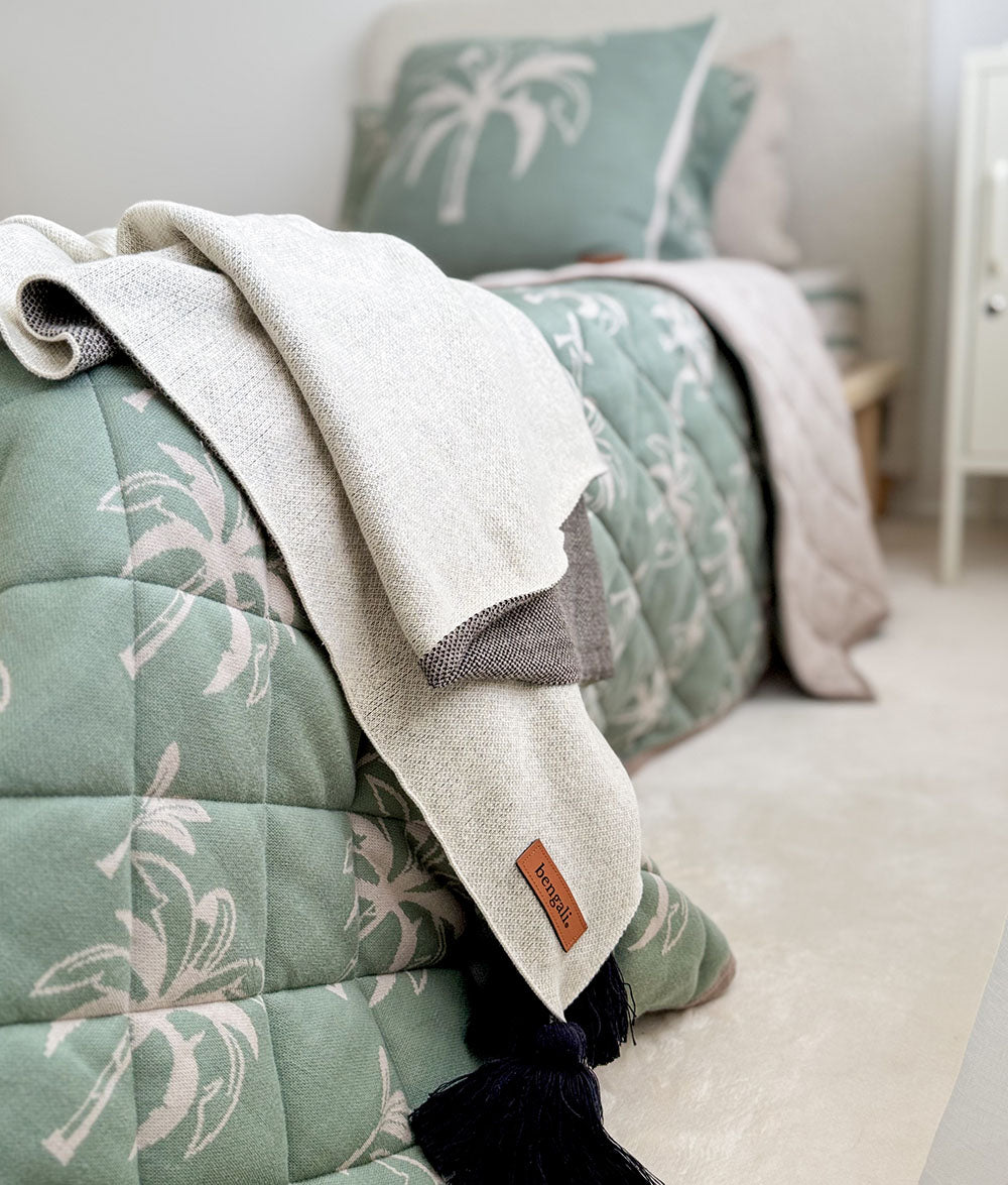 Green quilt with palm tree pattern and white blanket on a bed in a bedroom.