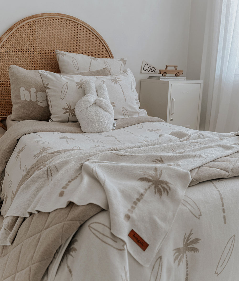 Neatly made bed with palm tree and surfboard patterned bedding, wicker headboard, and nightstand.