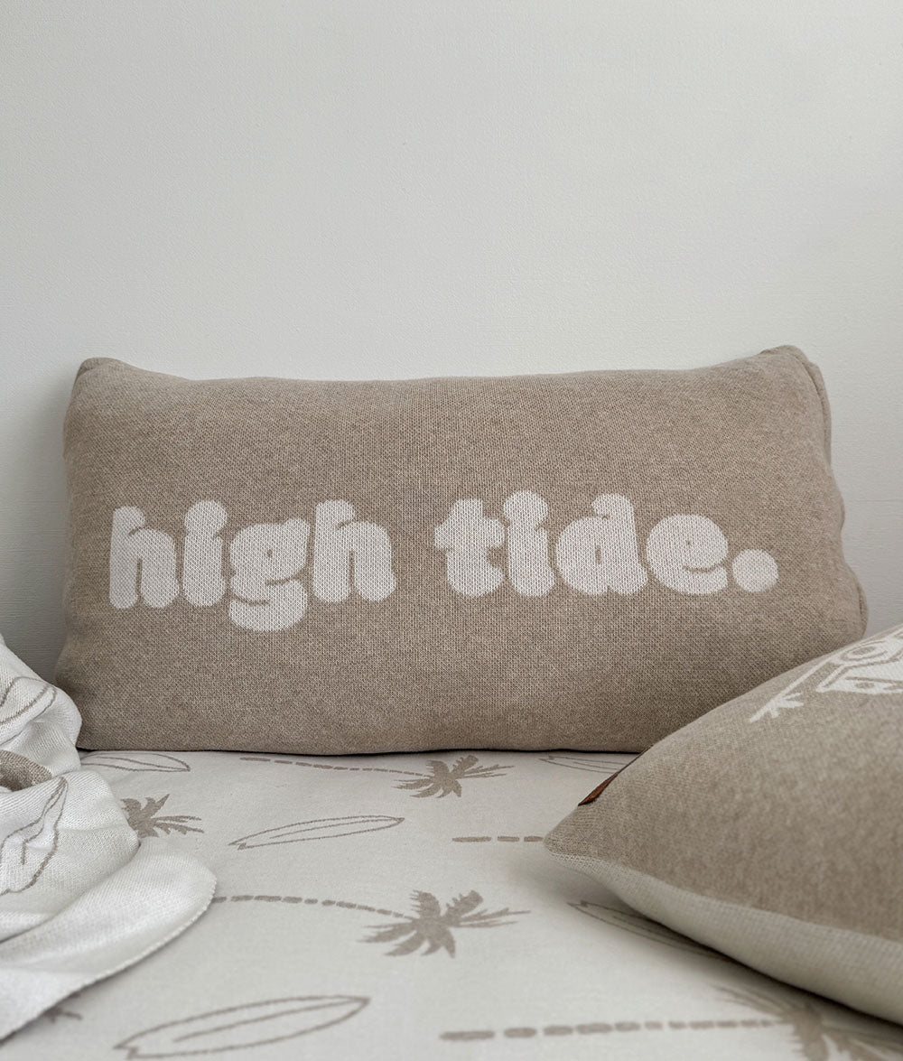 Beige pillow with 'high tide.' text on a bed with a white background
