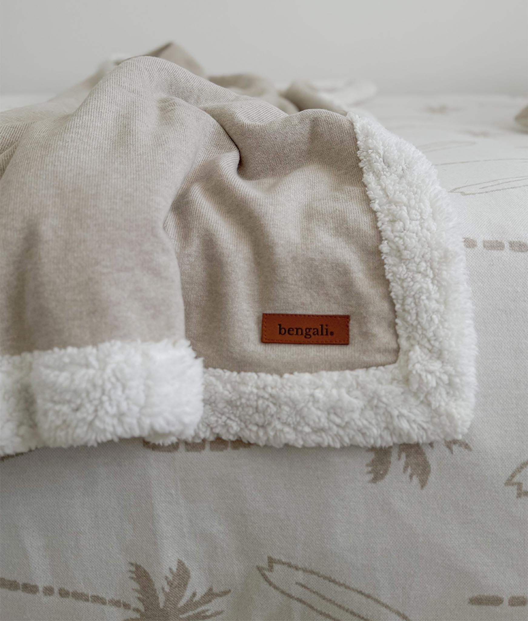Beige blanket with white sherpa lining and 'bengali' label on a bed with a patterned bedspread.