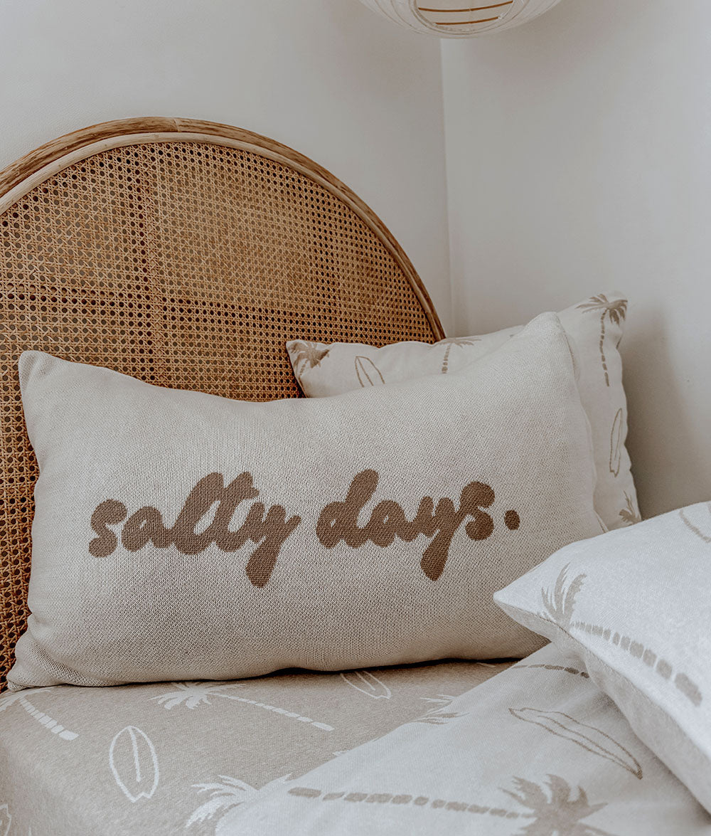 Beige pillow with 'salty days' text on a bed with wicker headboard