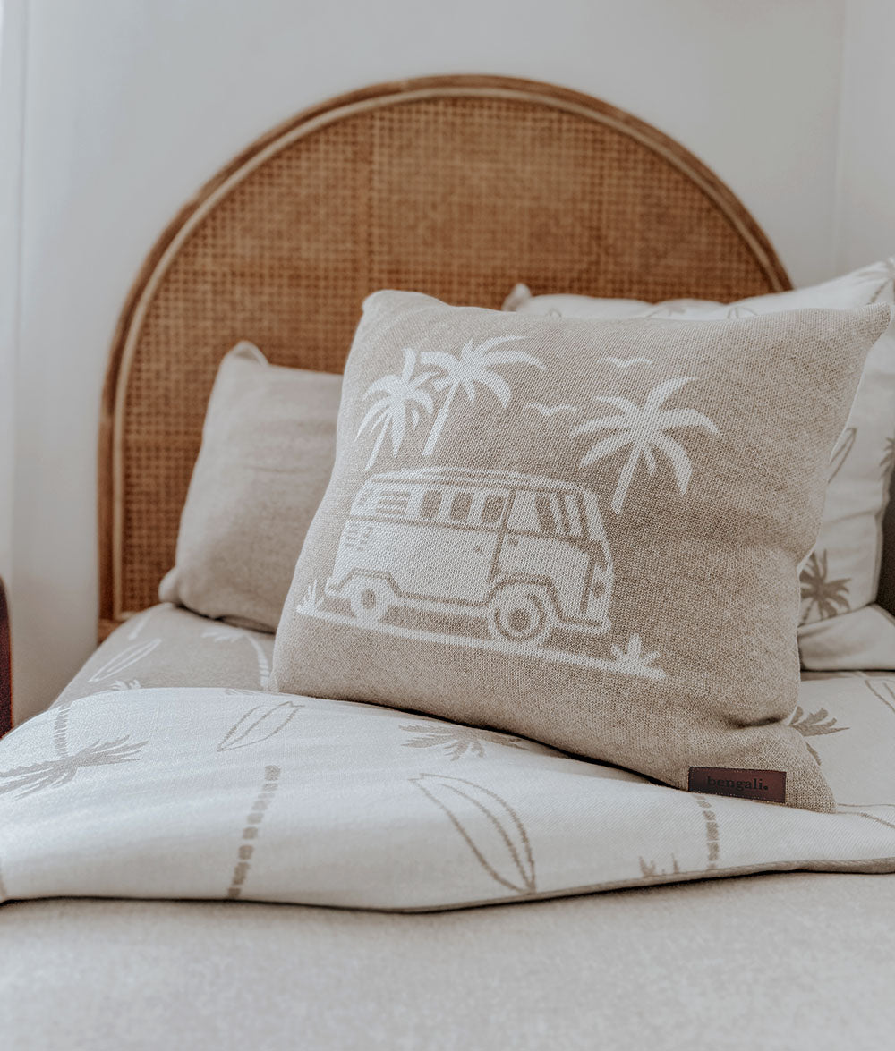 Decorative pillow with a van and palm tree design on a bed with a wicker headboard.