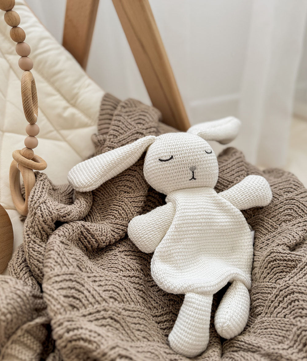 White knitted bunny toy on a textured blanket with a wooden baby gym in the background