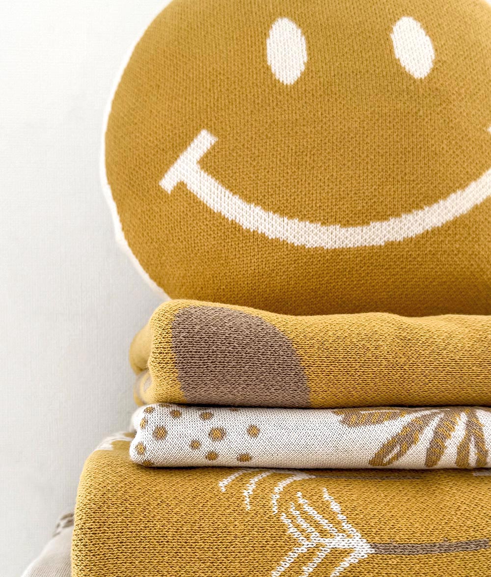 A stack of knitted baby blankets in warm mustard yellow and cream, featuring a large white smiley face on the top blanket and simple botanical and dot patterns on the folded layers below.