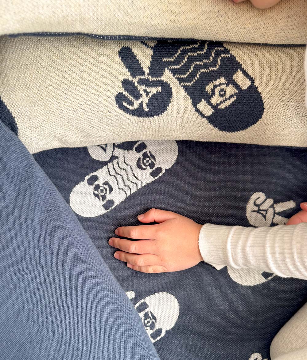 Close-up of the Skater Life reversible jersey cotton bedding, featuring navy and natural colourways with ivory skateboard and peace hand motifs. A child's hand rests on the soft knitted fabric, highlighting the reversible design and cosy texture.