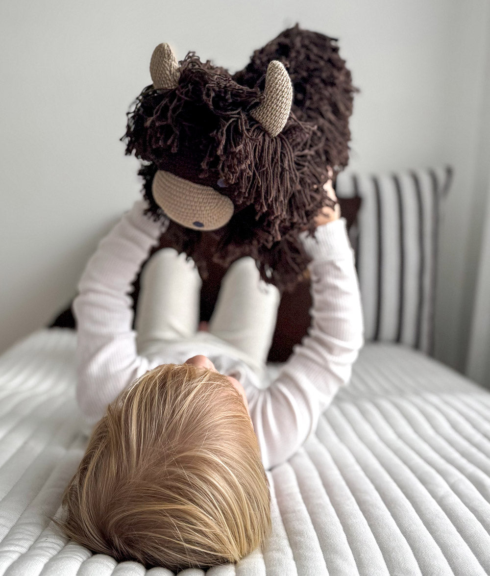 A child lying on a white bed holds a large dark brown highland cow toy with beige horns and a beige snout. The child is wearing a white long-sleeve top and white pants, with a striped pillow and a light gray wall in the background.