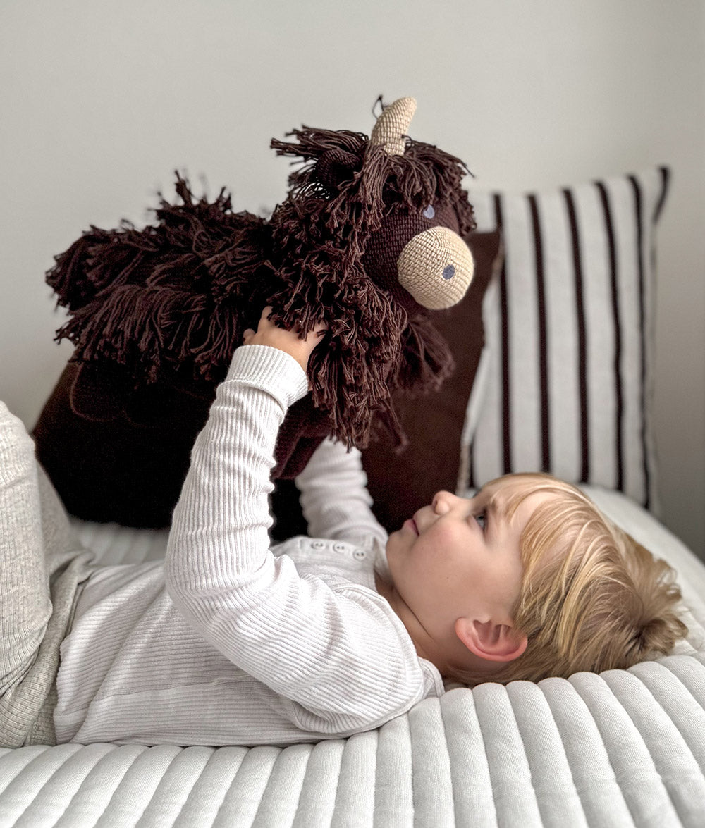 A young child lies on a ribbed white bed, holding a dark brown shaggy plush cow toy. The child is wearing a white ribbed long-sleeve top.
